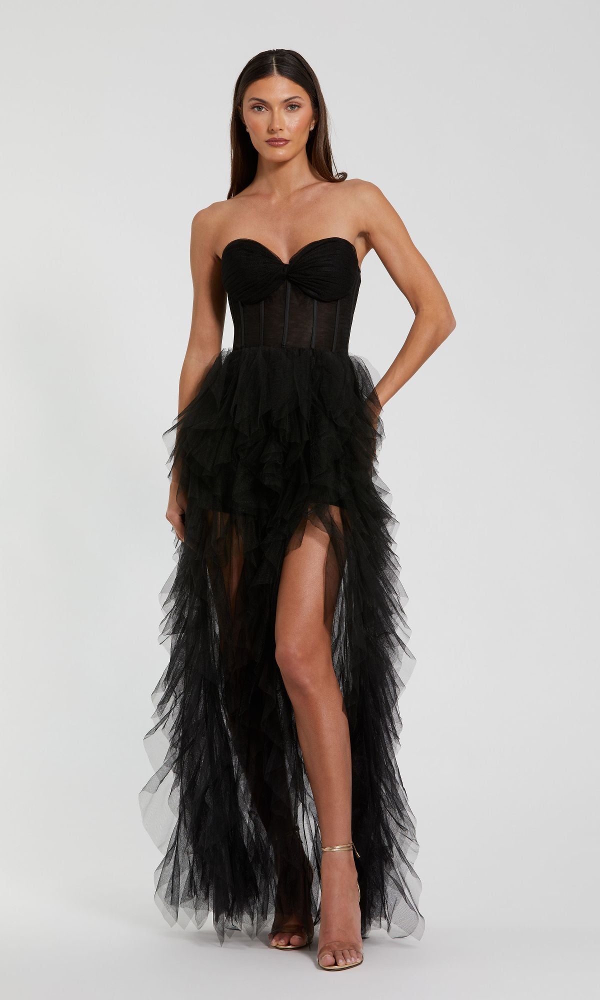 A woman poses against a plain background wearing the Mac Duggal 27532 Strapless Ruffled Long Prom Dress in black tulle, with a fitted bodice and a tiered, sheer high-low skirt.