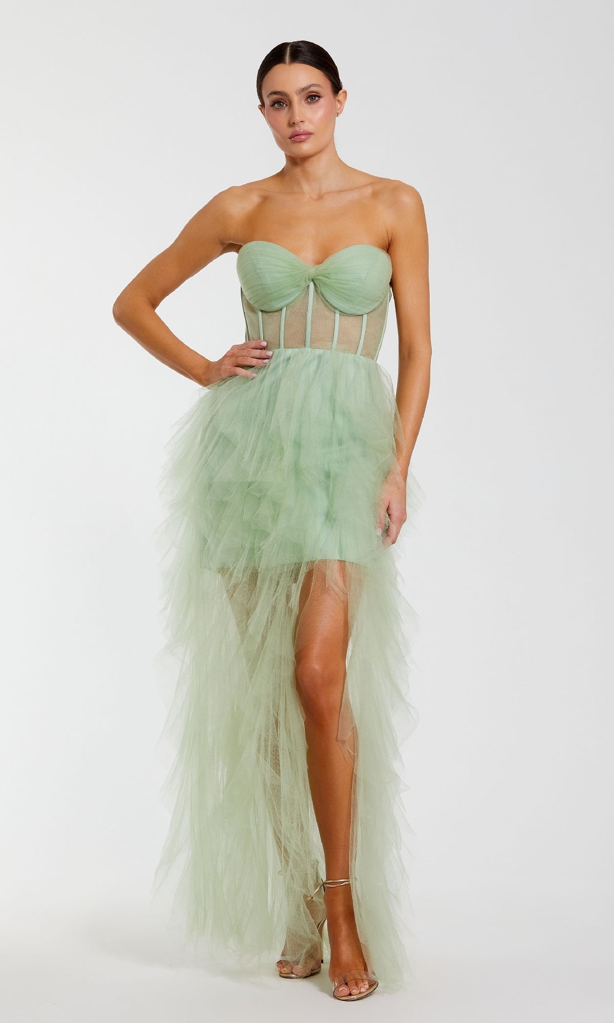 A woman models the Mac Duggal 27532 Strapless Ruffled Long Prom Dress in light green tulle, featuring a sheer corset bodice and an asymmetrical ruffled skirt, standing against a plain white background.