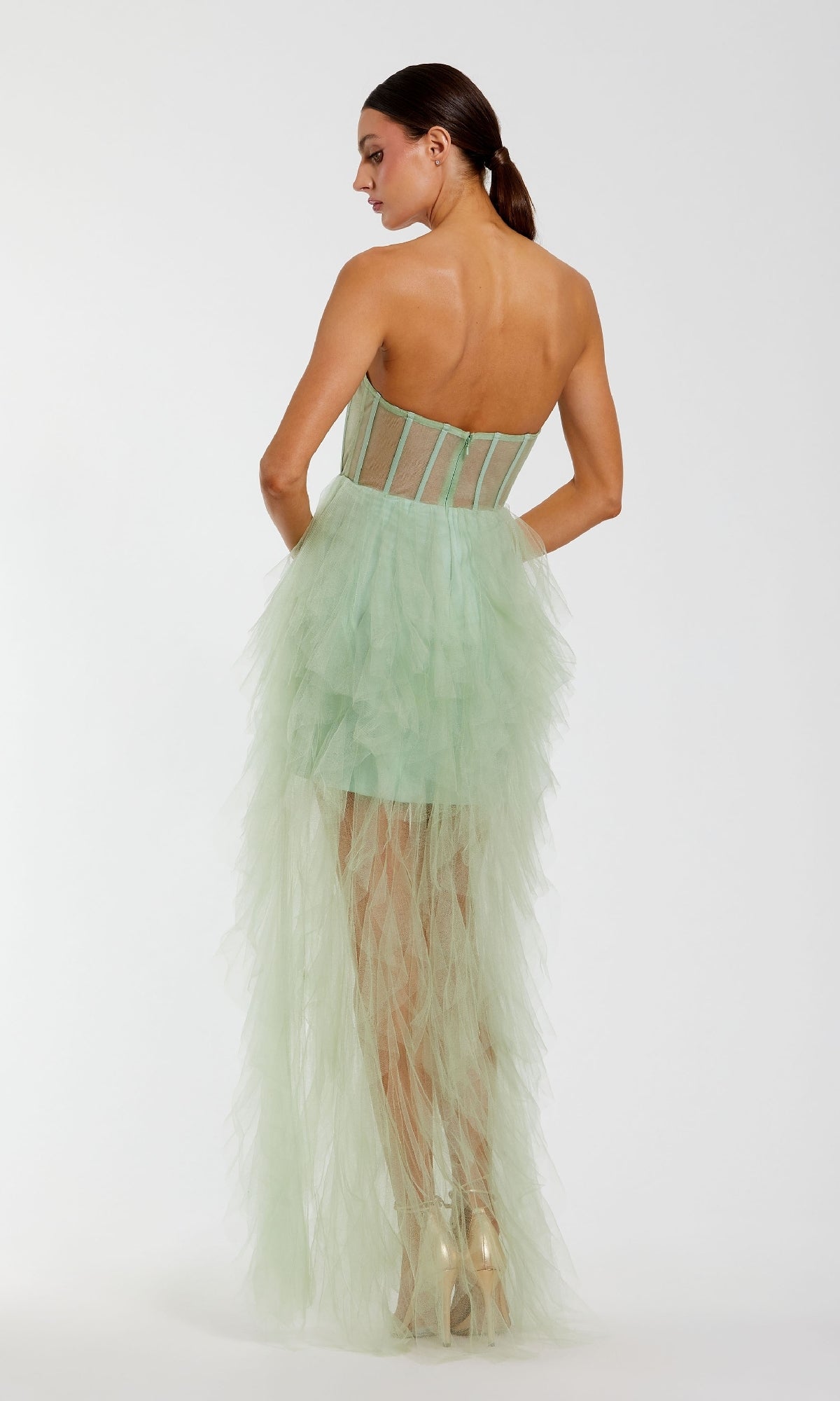 A woman faces away, wearing the Mac Duggal 27532 Strapless Ruffled Long Prom Dress in light green tulle with a sheer bodice and asymmetrical skirt, paired with nude heels.