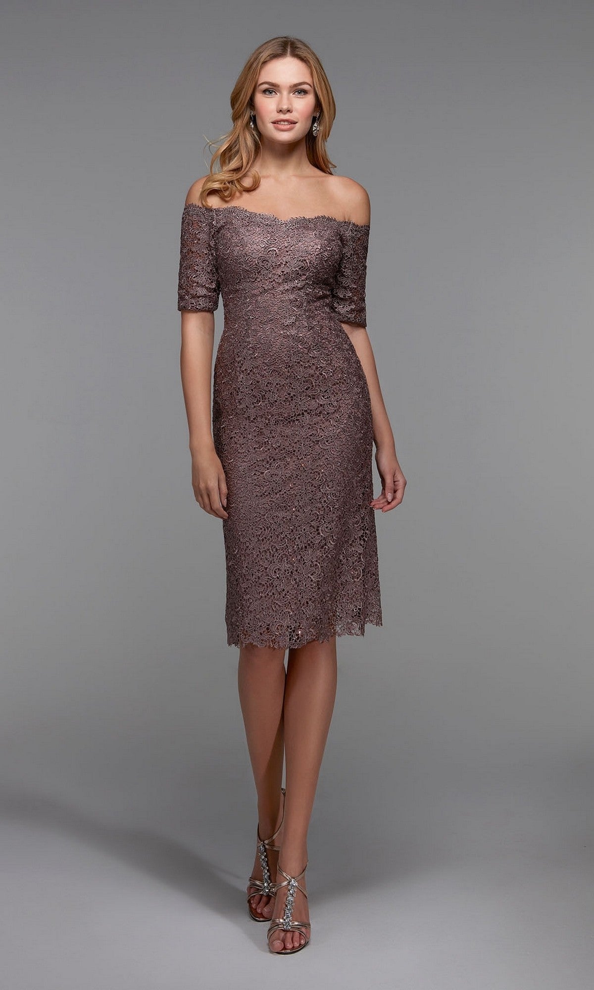 A woman wearing the Alyce 27535 Knee Length Semi-Formal Dress in Smoke, featuring an off-the-shoulder lace sheath design, paired with silver strappy heels, stands against a plain gray background.