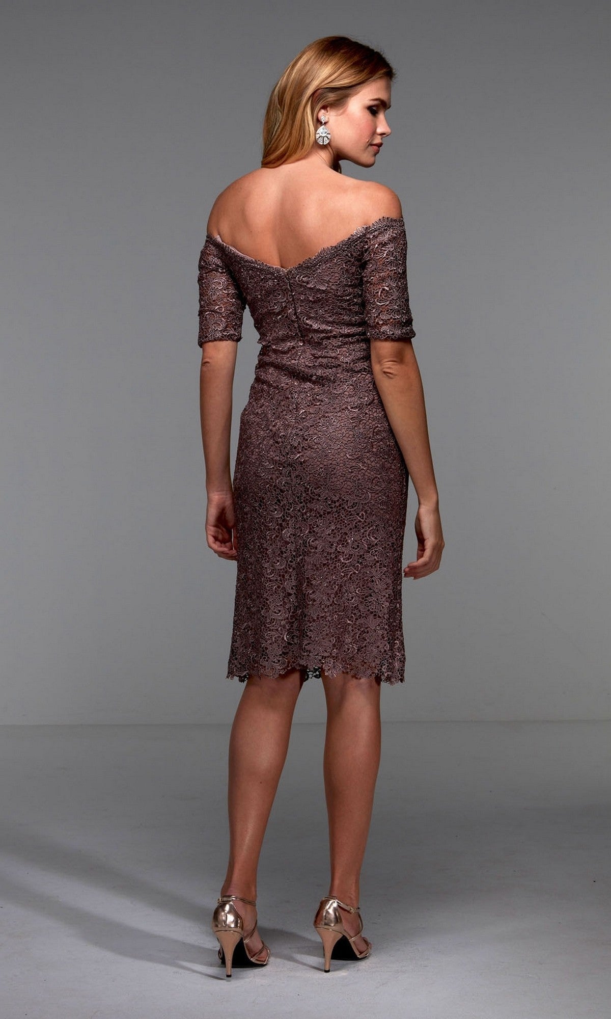A woman models the Alyce 27535, a knee-length semi-formal dress featuring an off-the-shoulder lace design in muted mauve, as she faces away to showcase the elegant back details paired with metallic heels.