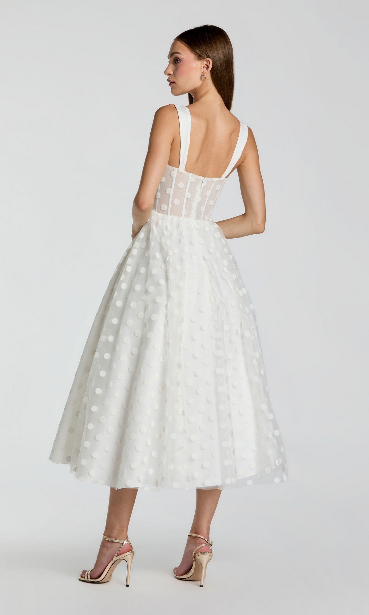 A woman seen from behind wears the Mac Duggal 27539 Black Midi Polka Dot Party Dress, featuring a sleeveless design, wide straps, tea-length sheer overlay, and paired with high-heeled sandals.
