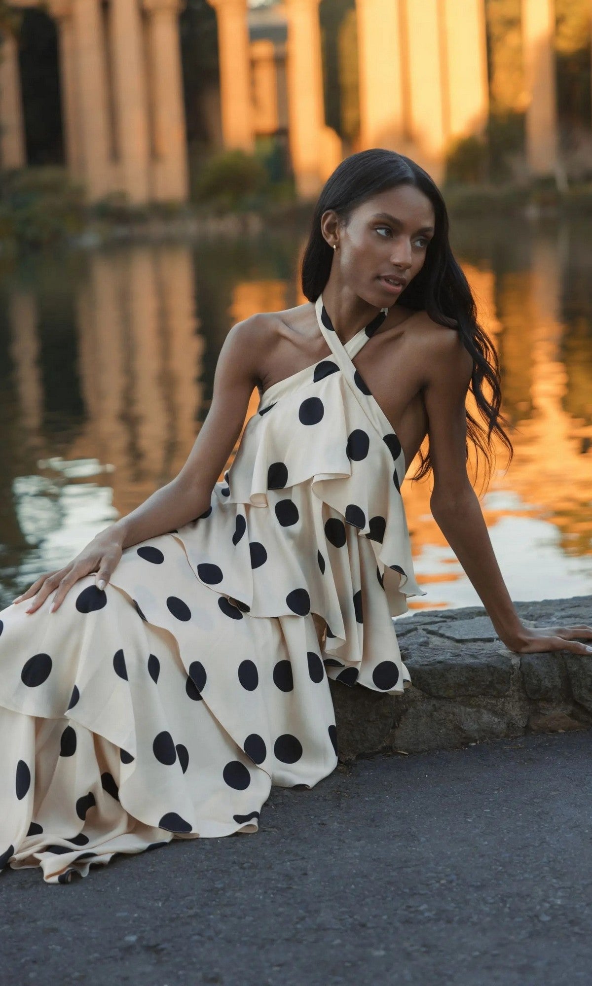 Woman sitting by a pond in a Long Formal Dress: Mac Duggal 27540, a cream, halter long dress with a large black polka-dot pattern and ruffled tiers.