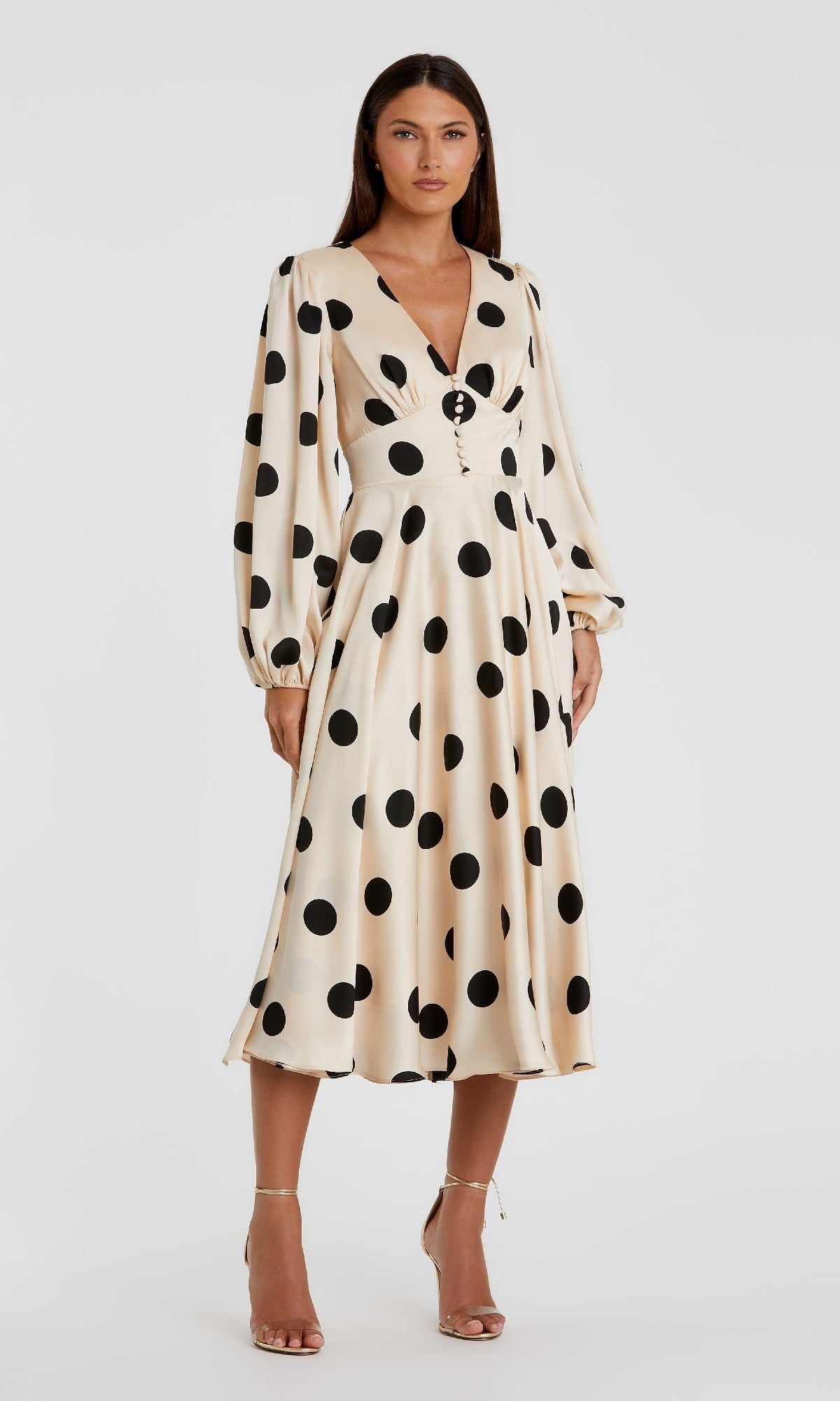 A woman wears the Mac Duggal 27542 Midi Polka-Dot Semi-Formal Dress, featuring cream fabric, long sleeves, a deep V-neckline, and button detail, styled with nude heels for a chic vintage-inspired party look.