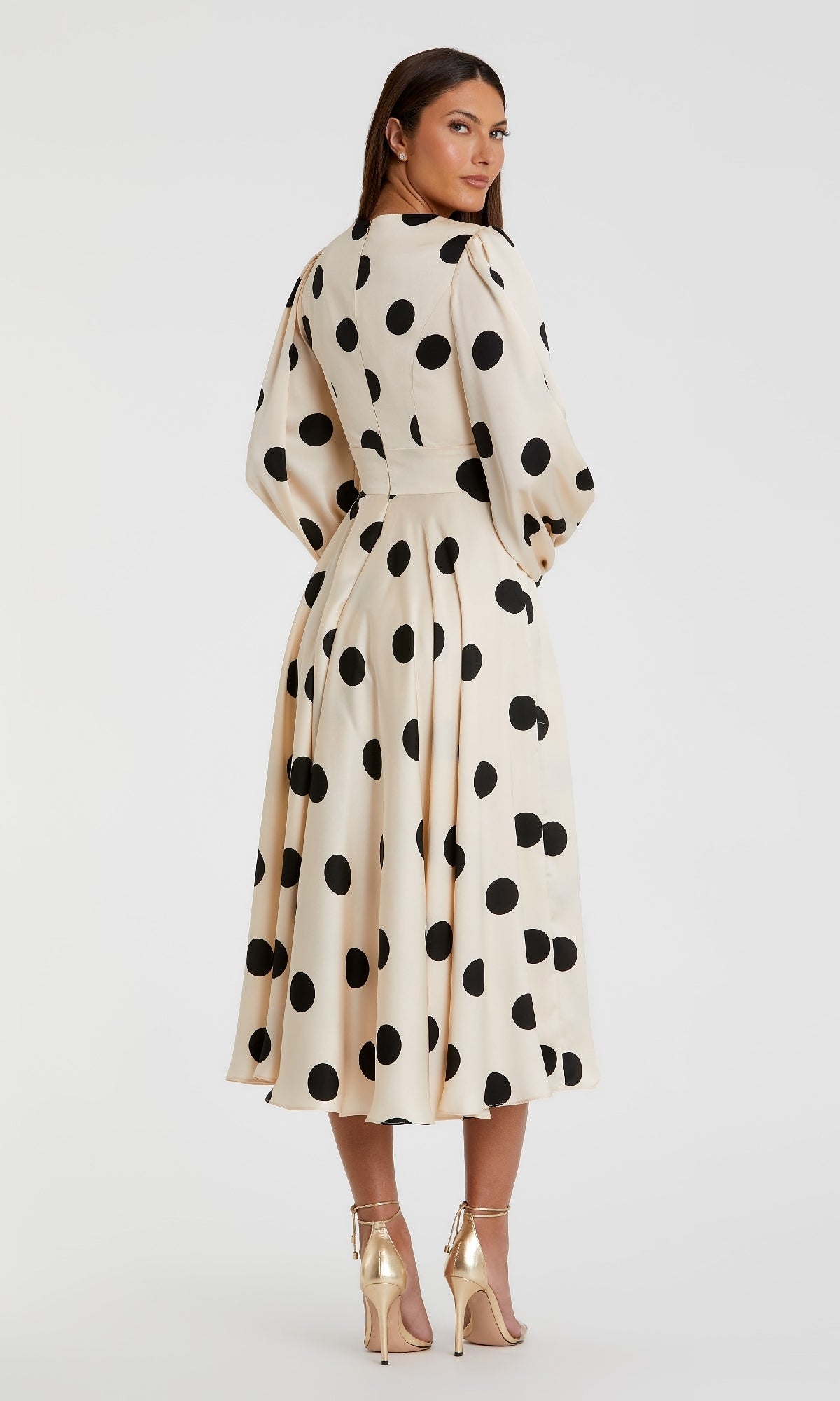 A woman stands with her back to the camera, modeling the Mac Duggal 27542: a cream-colored polka-dot semi-formal midi dress with long sleeves, paired with gold high-heeled sandals.