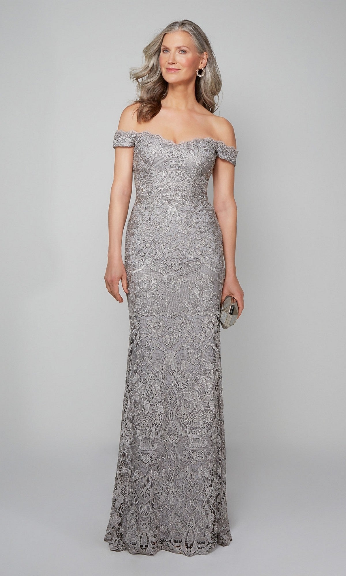 A woman in the Alyce 27553 off-the-shoulder, floor-length beaded lace formal dress holds a small clutch and poses elegantly against a plain light gray background.