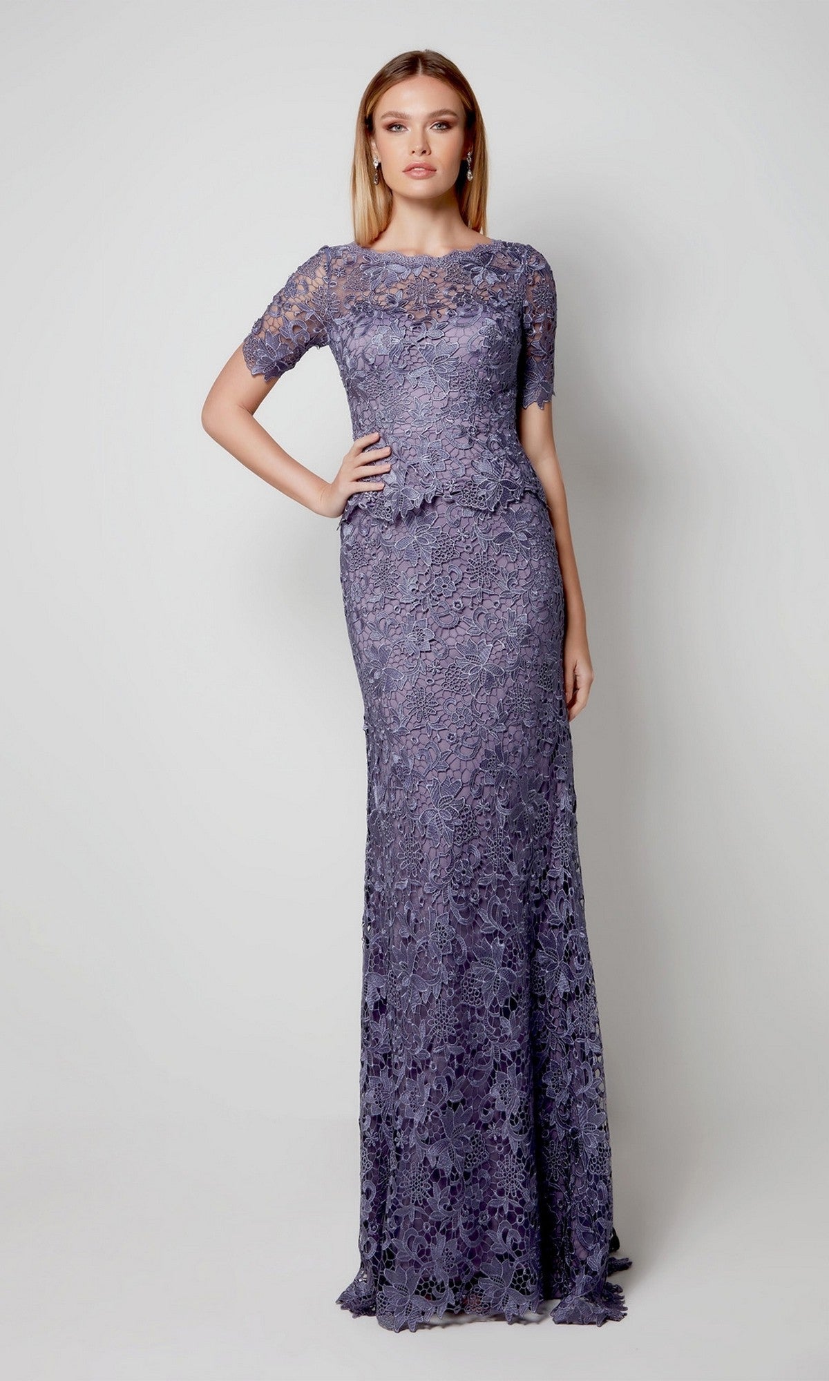 A woman poses against a plain backdrop wearing the Long Formal Dress: Alyce 27555 in Storm Cloud—a fitted, floor-length lace gown with short sleeves and a scalloped neckline, ideal for an elegant mother-of-the-bride look.