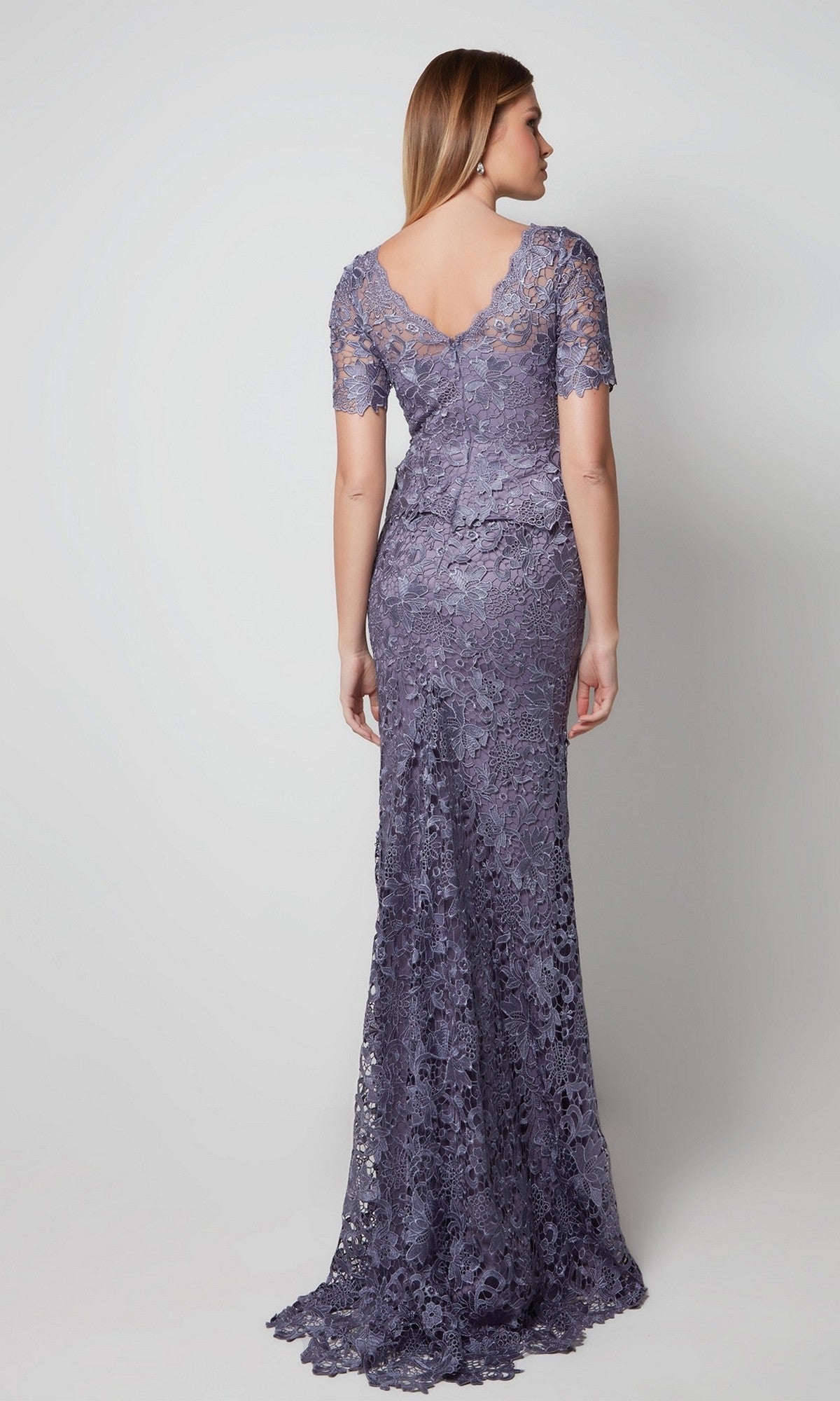 A woman stands with her back to the camera in a floor-length, short-sleeved lavender lace Alyce 27555 formal dress against a plain background.
