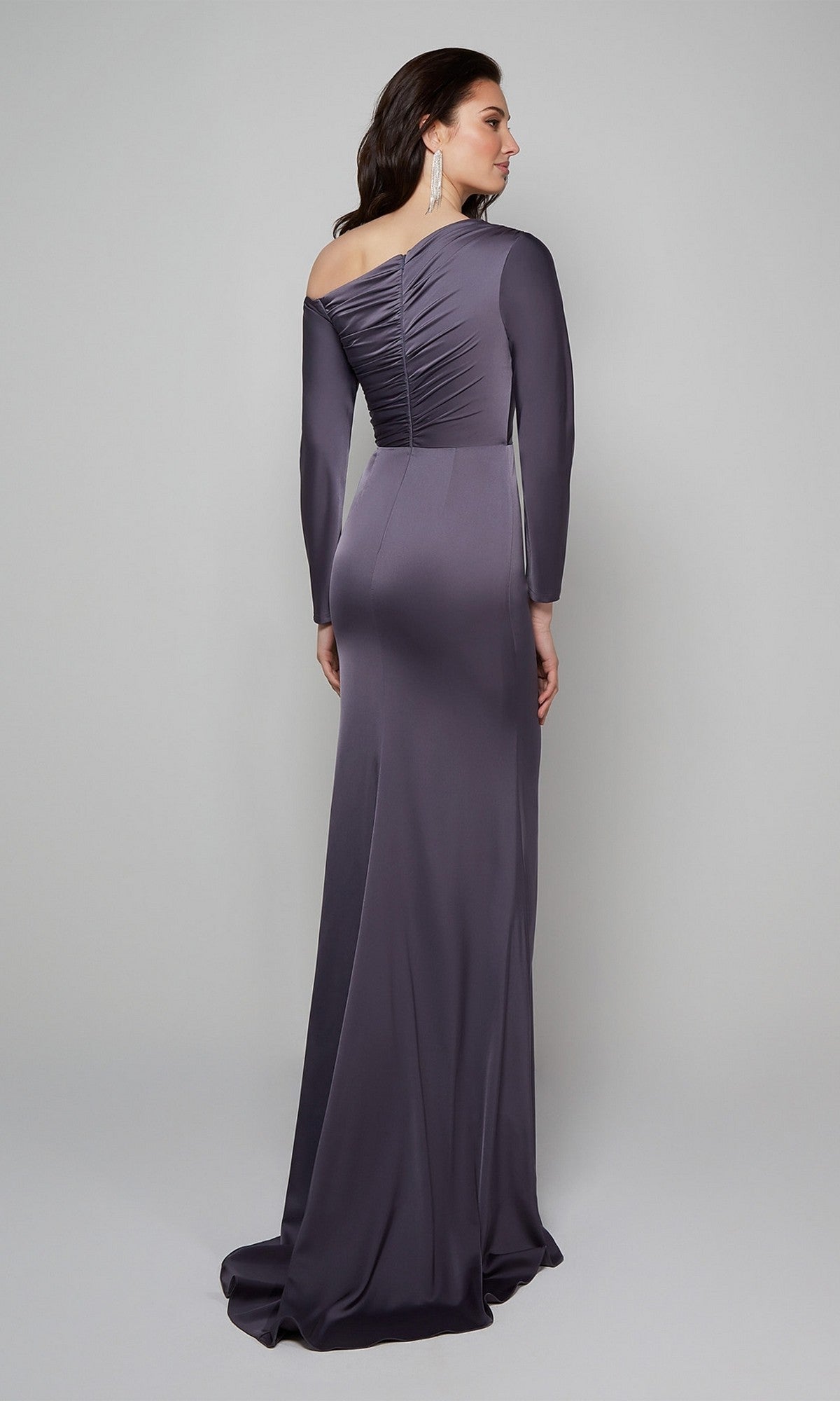 Woman wearing the Alyce 27557, a long, fitted dark purple formal dress with long sleeves and ruched back detail, standing against a light gray background. An elegant choice for a stylish wedding guest look.