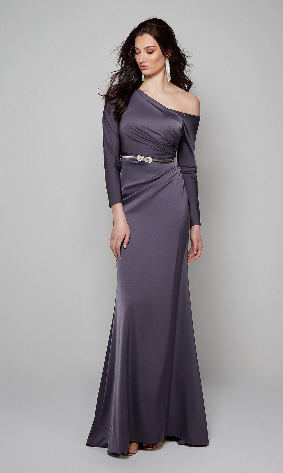 Wearing the Alyce 27557 long formal dress in slate gray satin, featuring a one-shoulder detail, long sleeves, and a decorative belt with drop earrings, she stands poised—an elegant choice for any wedding guest.