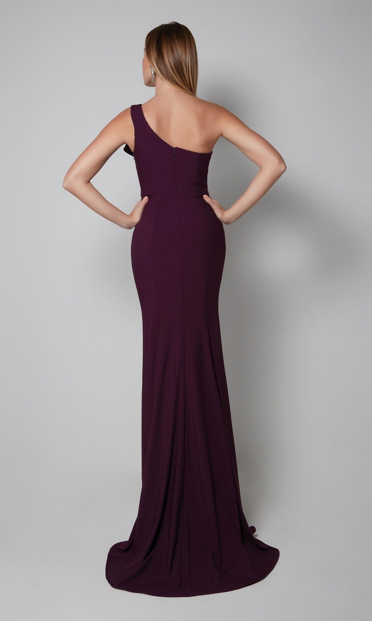 A woman, hands on hips, faces away in the Alyce 27575 long formal dress—an elegant, amethyst, floor-length gown with a one-shoulder design—set against a plain gray background.