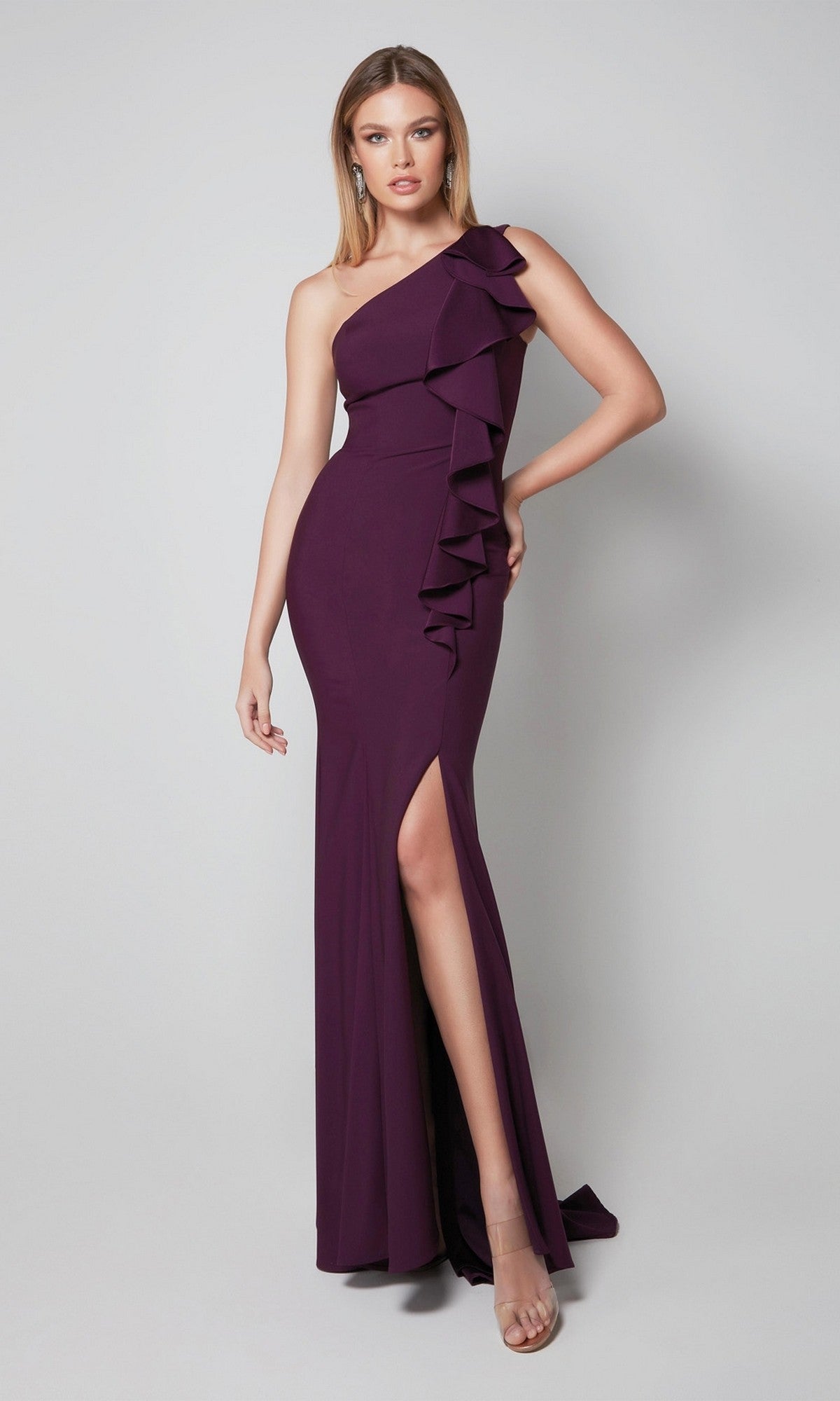 A woman stands against a plain light background wearing the Alyce 27575, a floor-length, one-shoulder amethyst formal dress featuring ruffle detailing and a thigh-high slit.