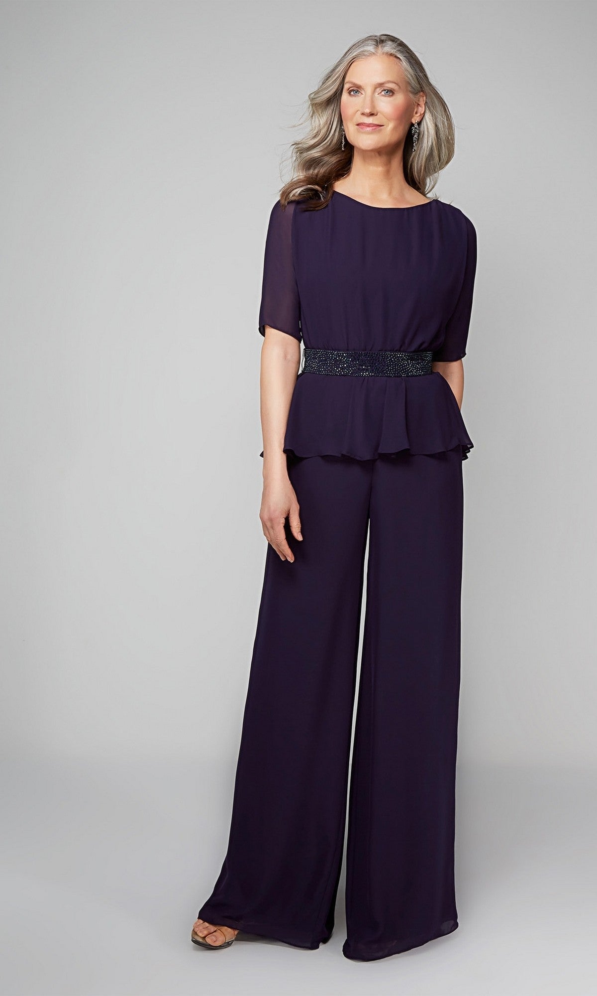 A woman with long gray hair wears the Alyce 27576 Long Formal Jumpsuit in Patriot Blue, featuring short sleeves and wide-leg pants, paired with open-toe shoes, standing against a plain light gray background.