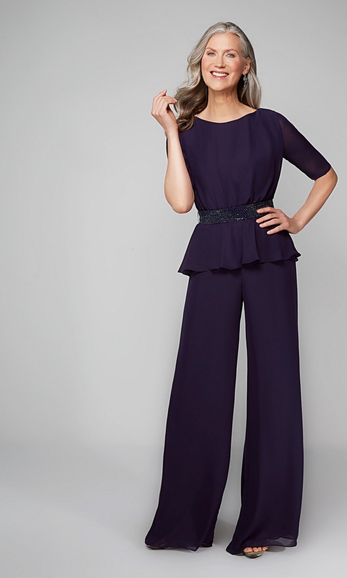 A smiling woman with long gray hair wears the Alyce 27576 long formal jumpsuit, featuring short sleeves and a beaded waist detail, standing against a plain light background.