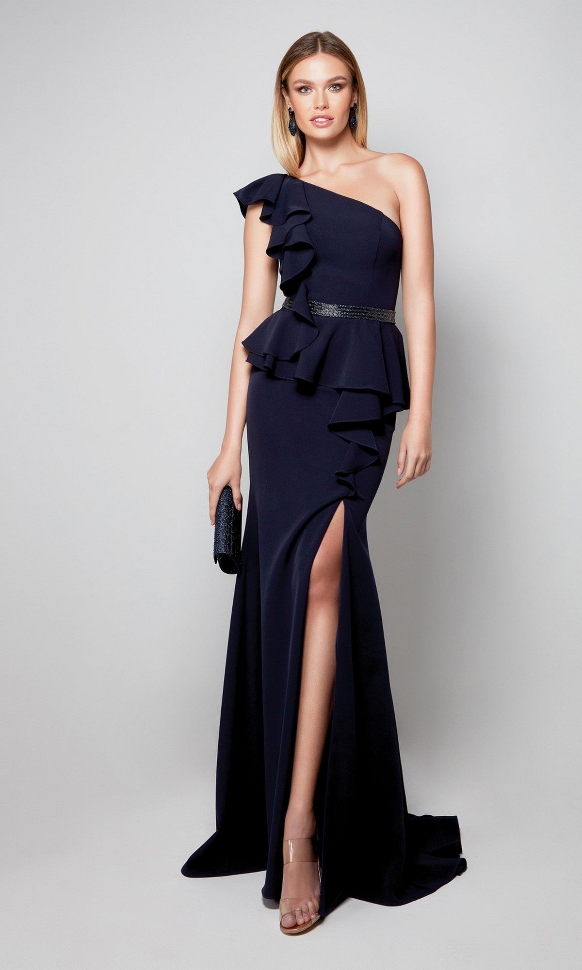 A woman in a midnight Alyce 27577 long formal dress—one-shoulder with ruffle detail, beaded belt, thigh-high slit—paired with clear heels and a black clutch, stands against a plain background.