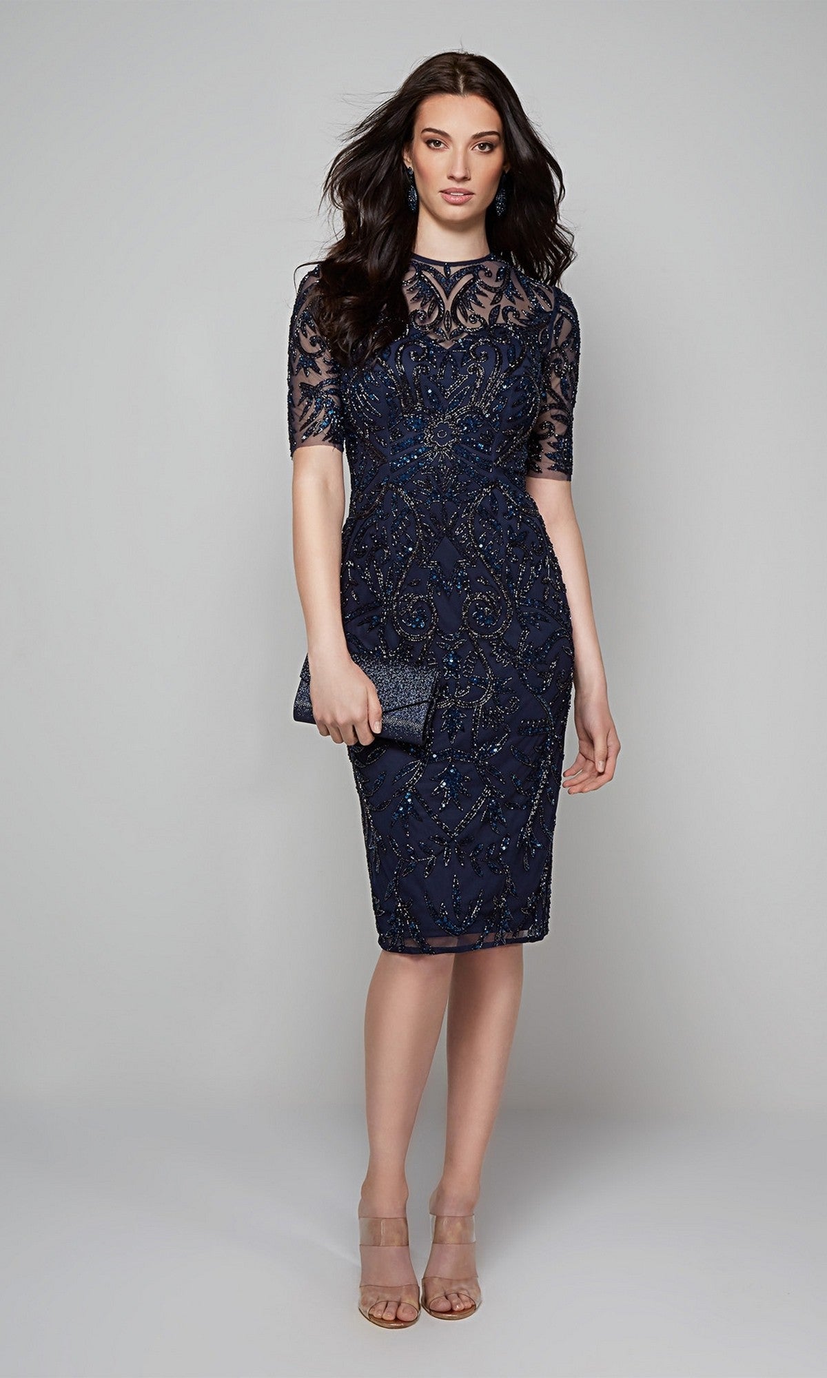 A woman in the Alyce 27583 knee-length semi-formal dress with sheer sleeves and embellishments, paired with a matching clutch and nude heels, poses against a plain light gray background.