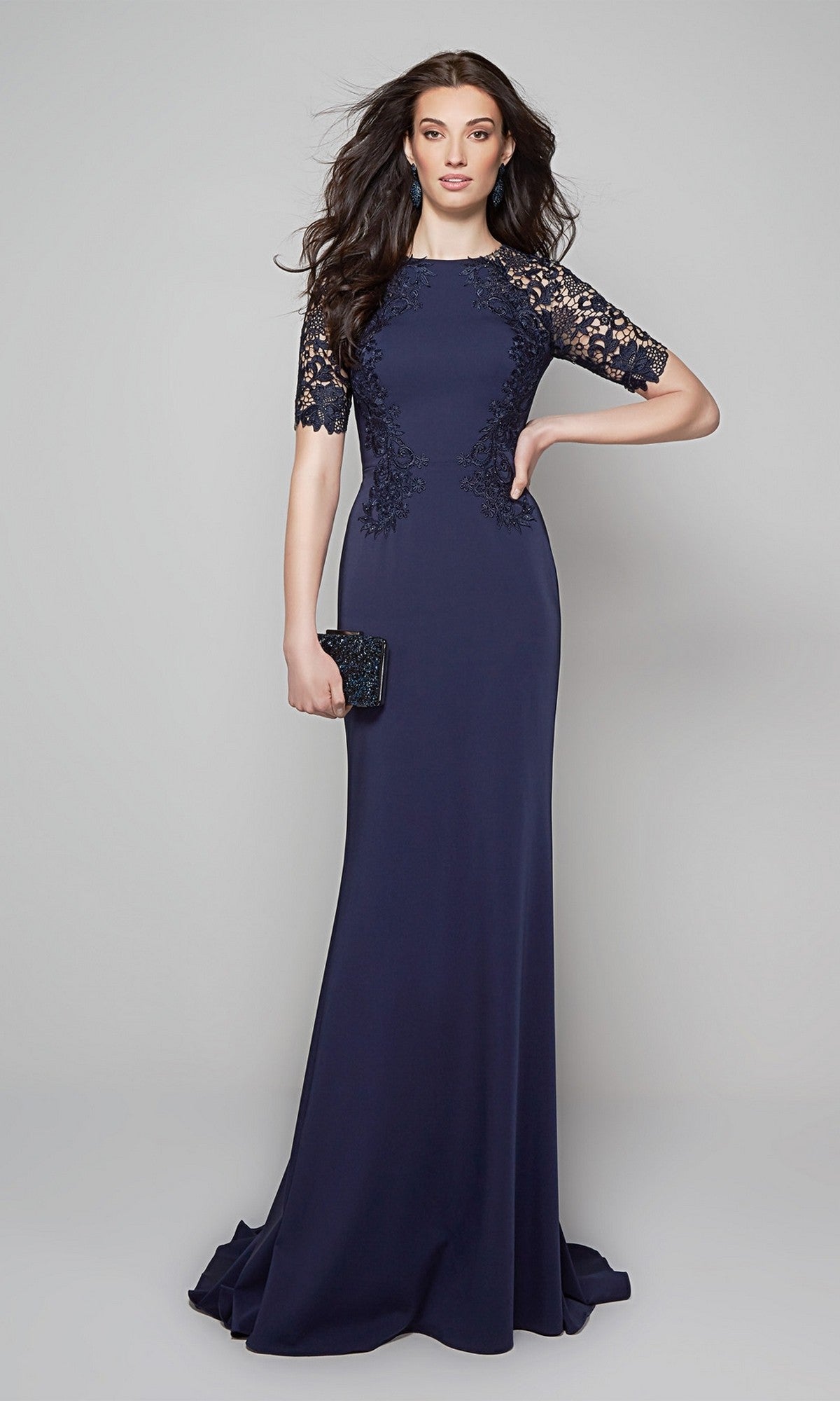 A woman in the Alyce 27585 long formal dress in midnight, featuring a lace bodice and lace-detailed sleeves, holds a black clutch while standing against a plain gray background.