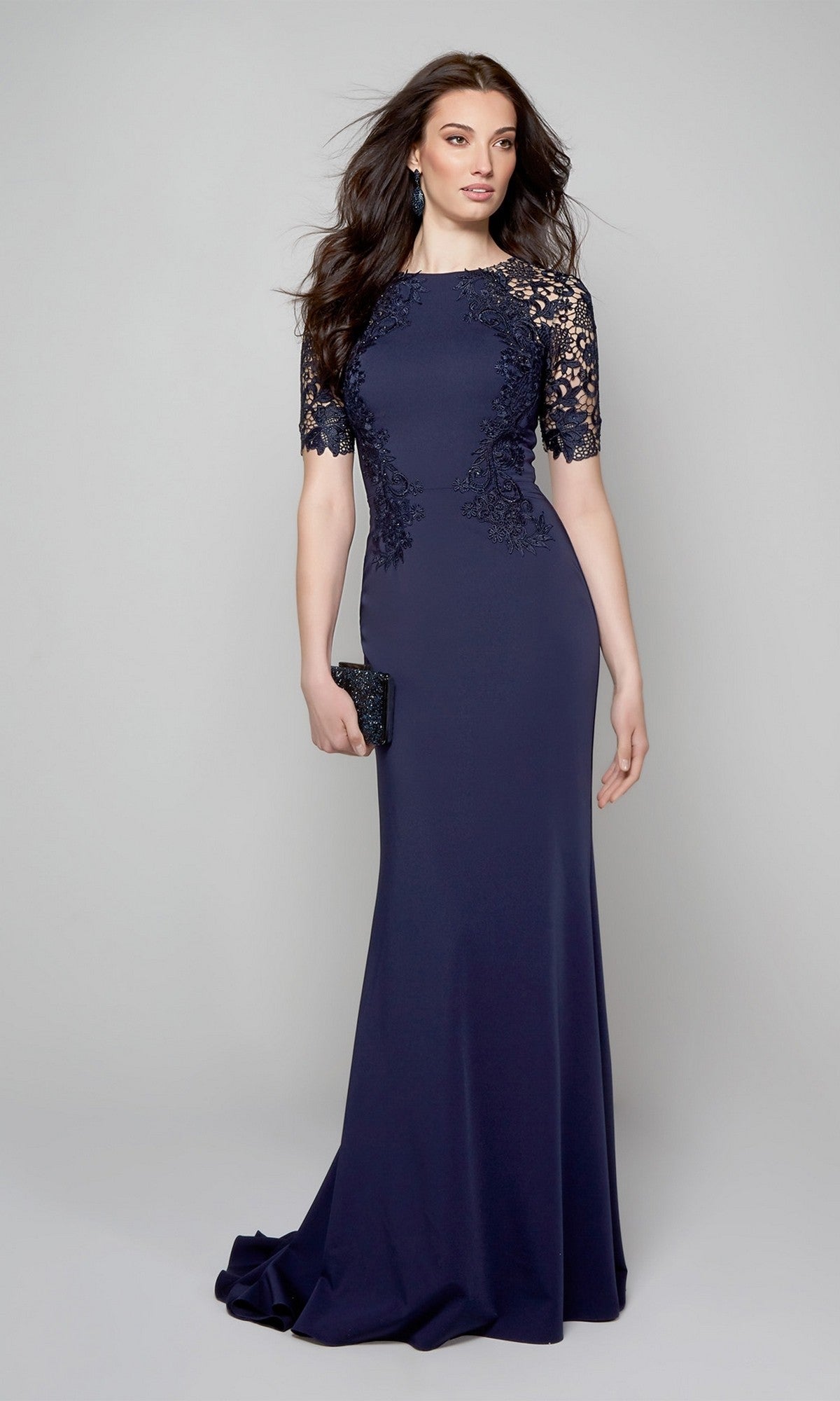 A woman wearing the Alyce 27585 long formal dress with a lace bodice and sleeves holds a black clutch, standing against a plain light gray background.