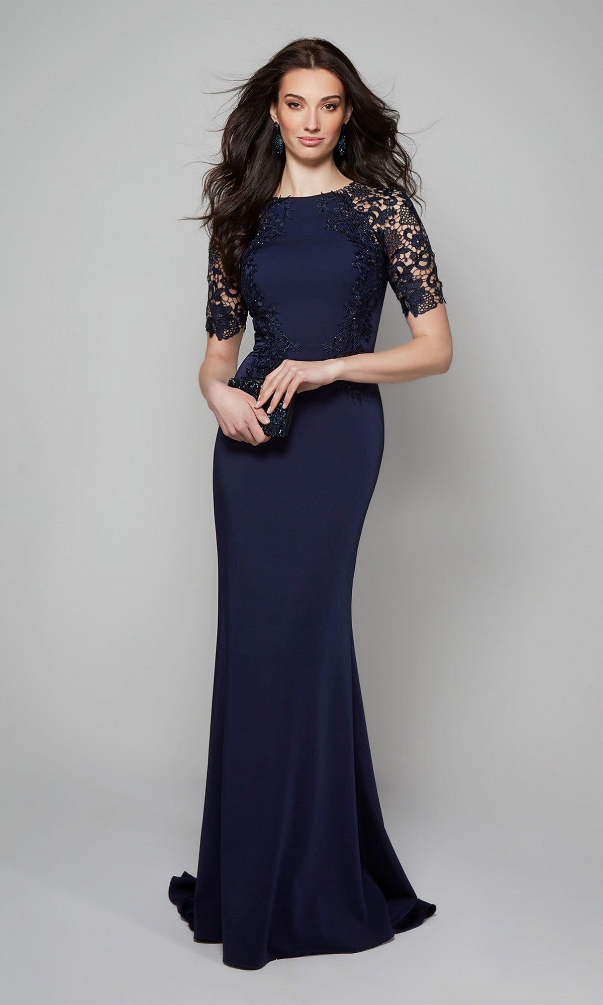 A woman models the Alyce 27585, a navy blue floor-length sheath formal dress with lace sleeves, holding a small black clutch against a light gray background.