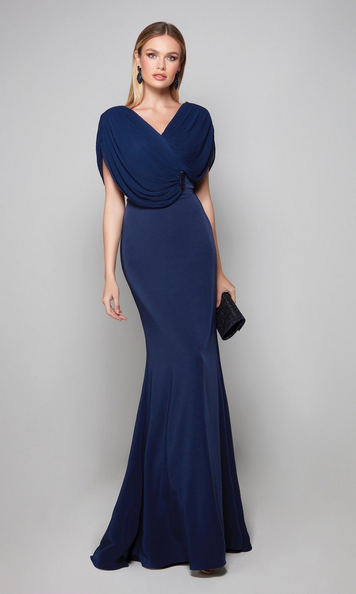 A woman wears the Alyce 27591 long formal dress in midnight, featuring a fitted crepe silhouette and draped shoulders, holding a black clutch against a plain light gray background.