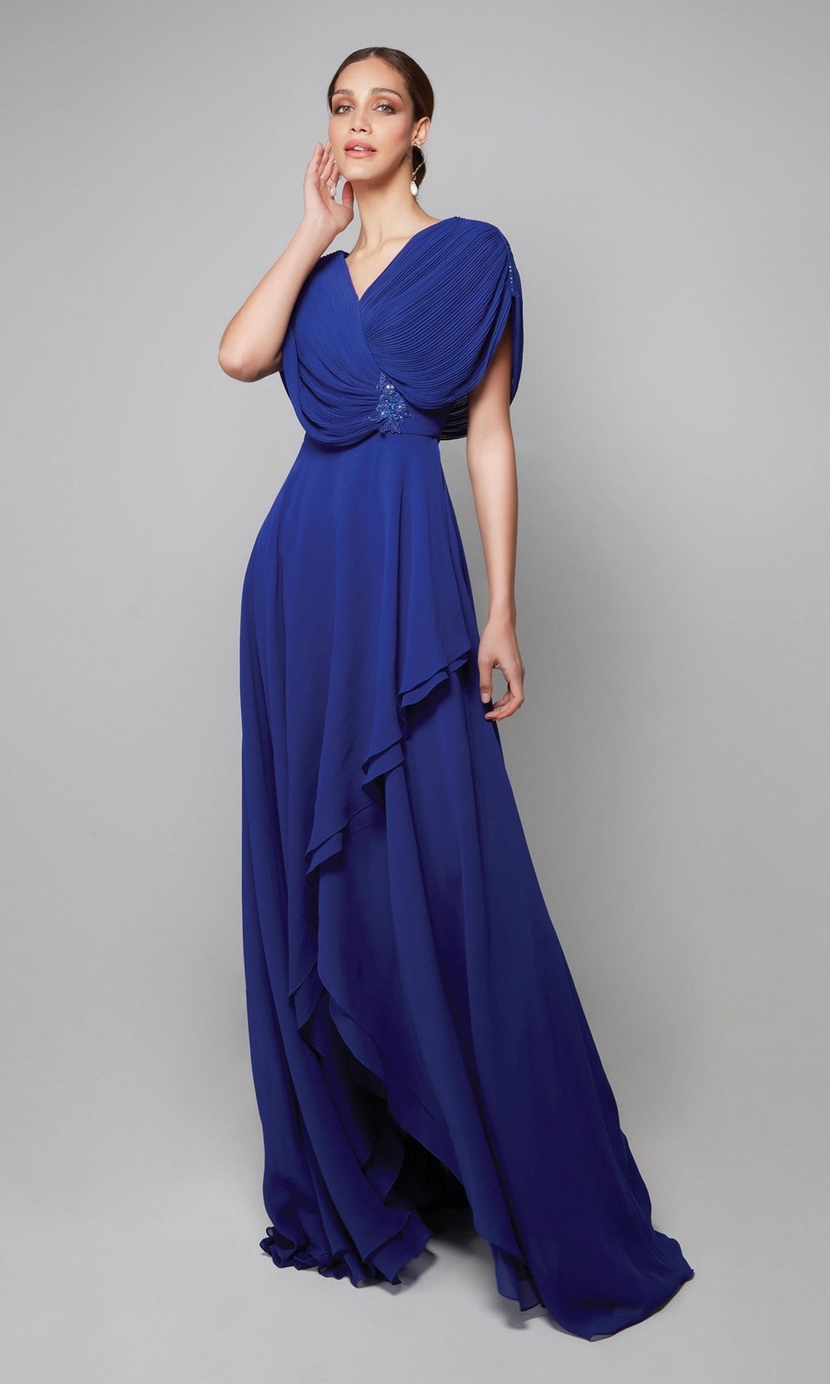 A woman poses against a plain gray background wearing the Alyce 27592 long formal dress in cobalt, featuring layered draped fabric, cape sleeves, and a fitted waist.
