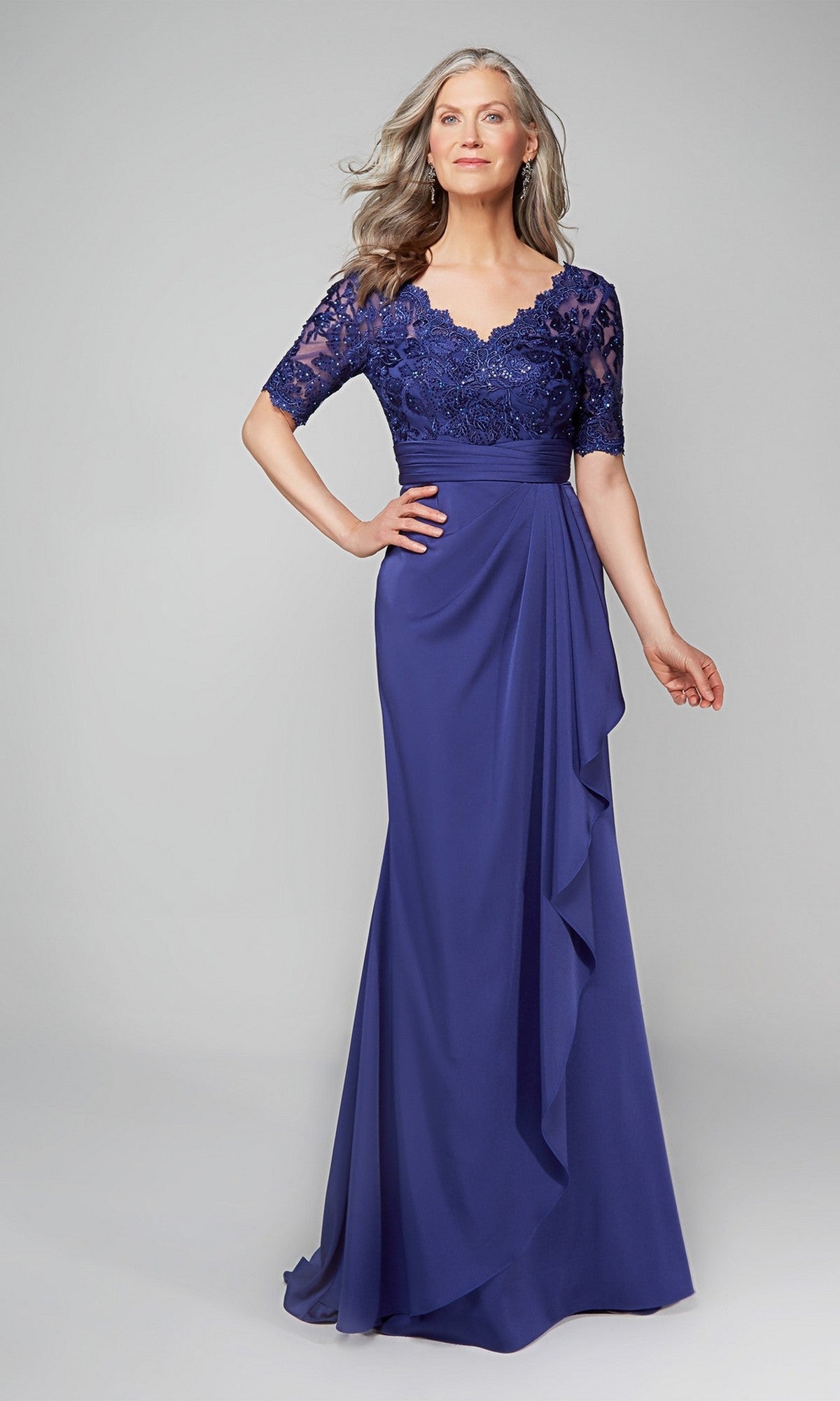 A woman wearing the Alyce 27595 long formal dress in navy, featuring a lace bodice, short sleeves, and floral appliqué details, stands against a plain light gray background.