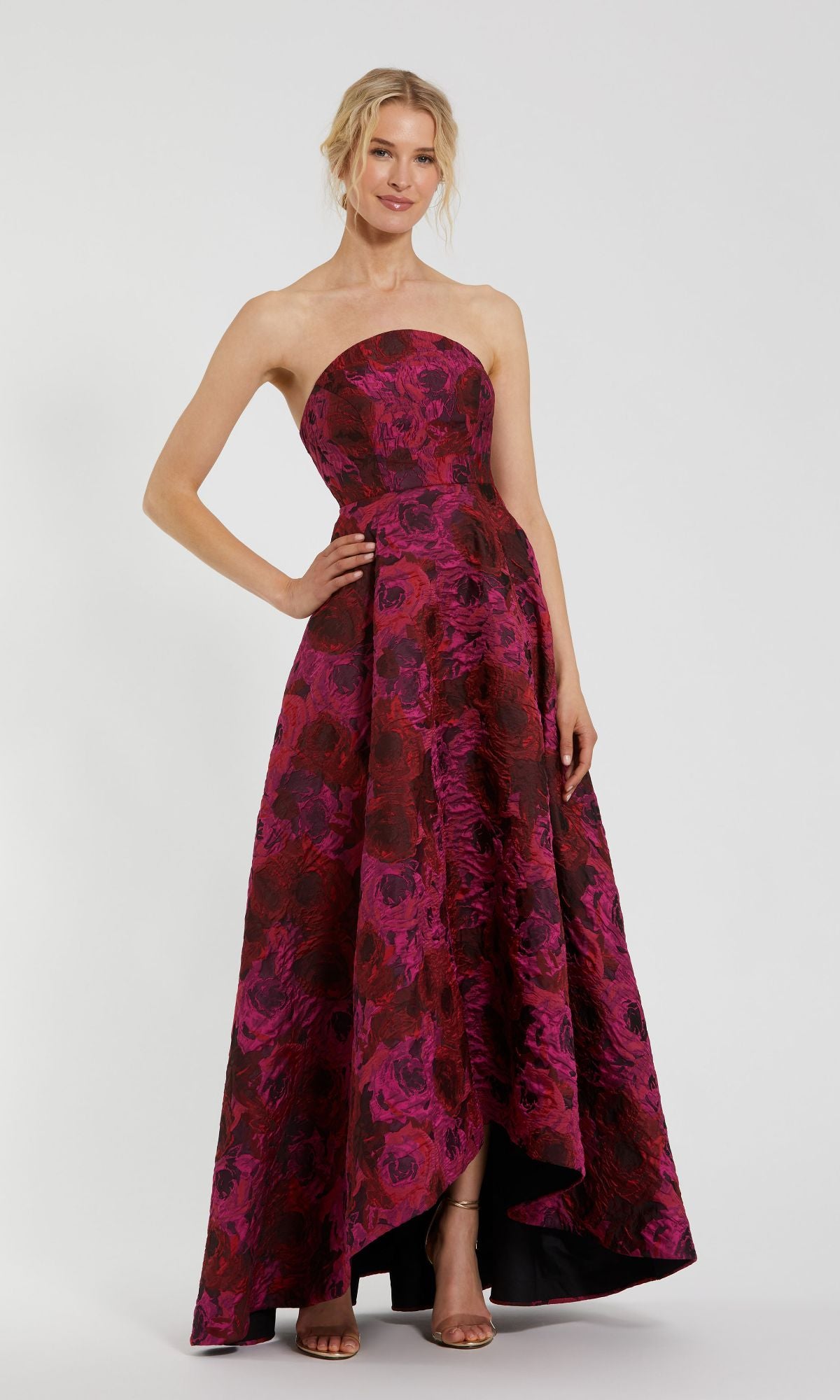 A woman poses against a plain background wearing the Pink Floral Formal Evening Gown, Mac Duggal 27596—a floor-length, strapless dress with a brocade floral pattern.