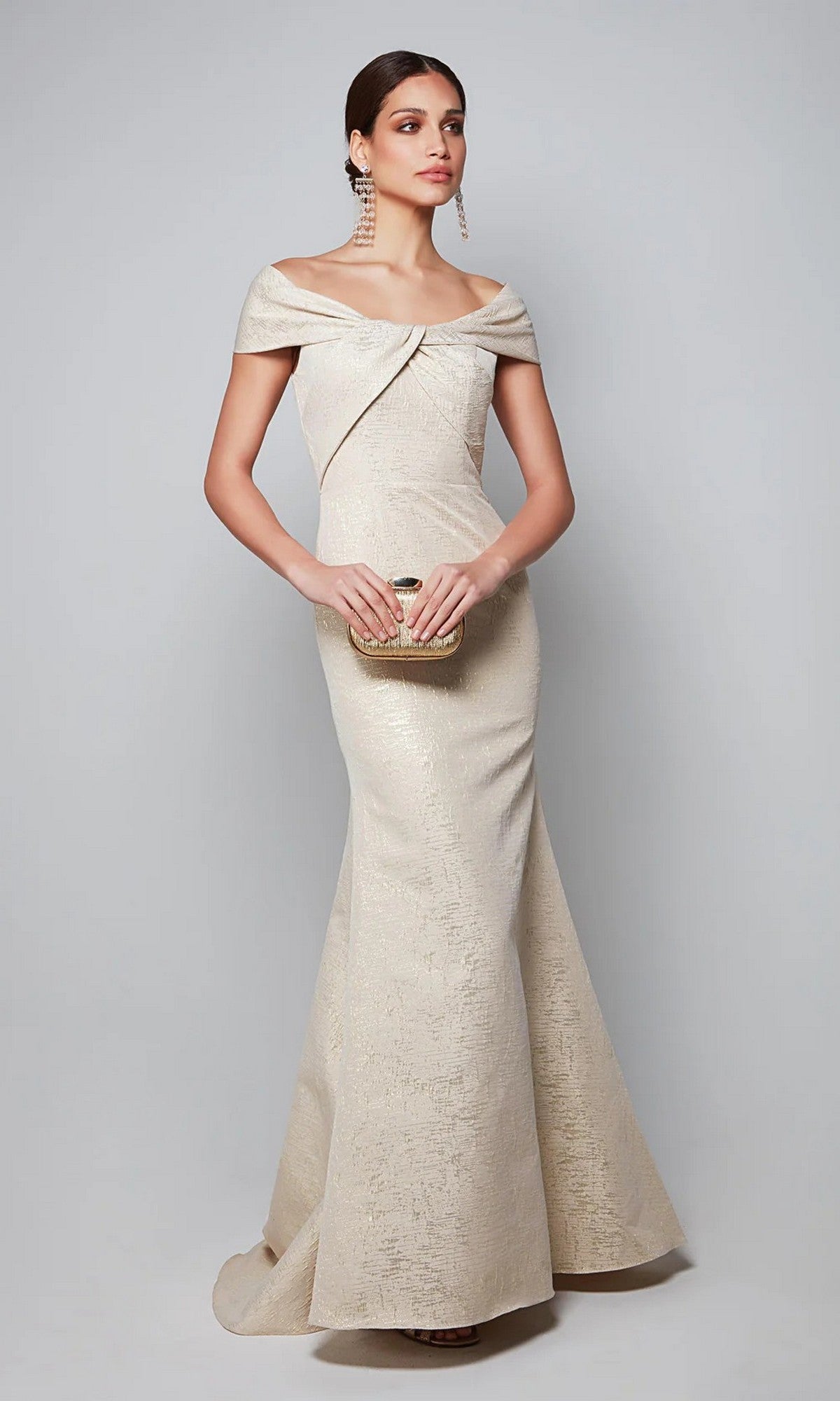 A woman in the Alyce 27613 long formal dress, sterling/gold color, holds a small clutch and stands gracefully against a plain light gray background.