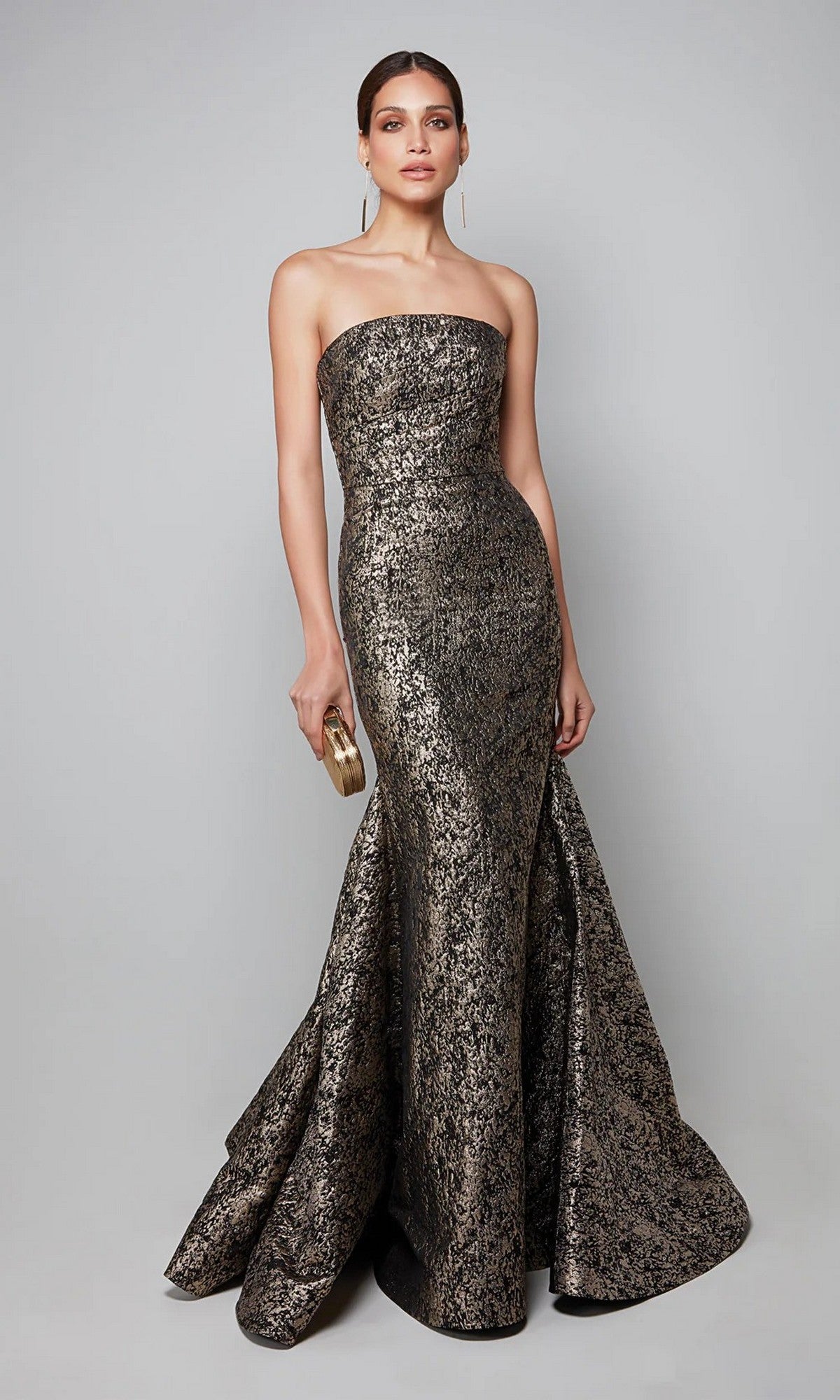 A woman in the Alyce 27621 long formal dress in gold/black, featuring a strapless jacquard mermaid silhouette, holds a small clutch and poses against a plain gray background.