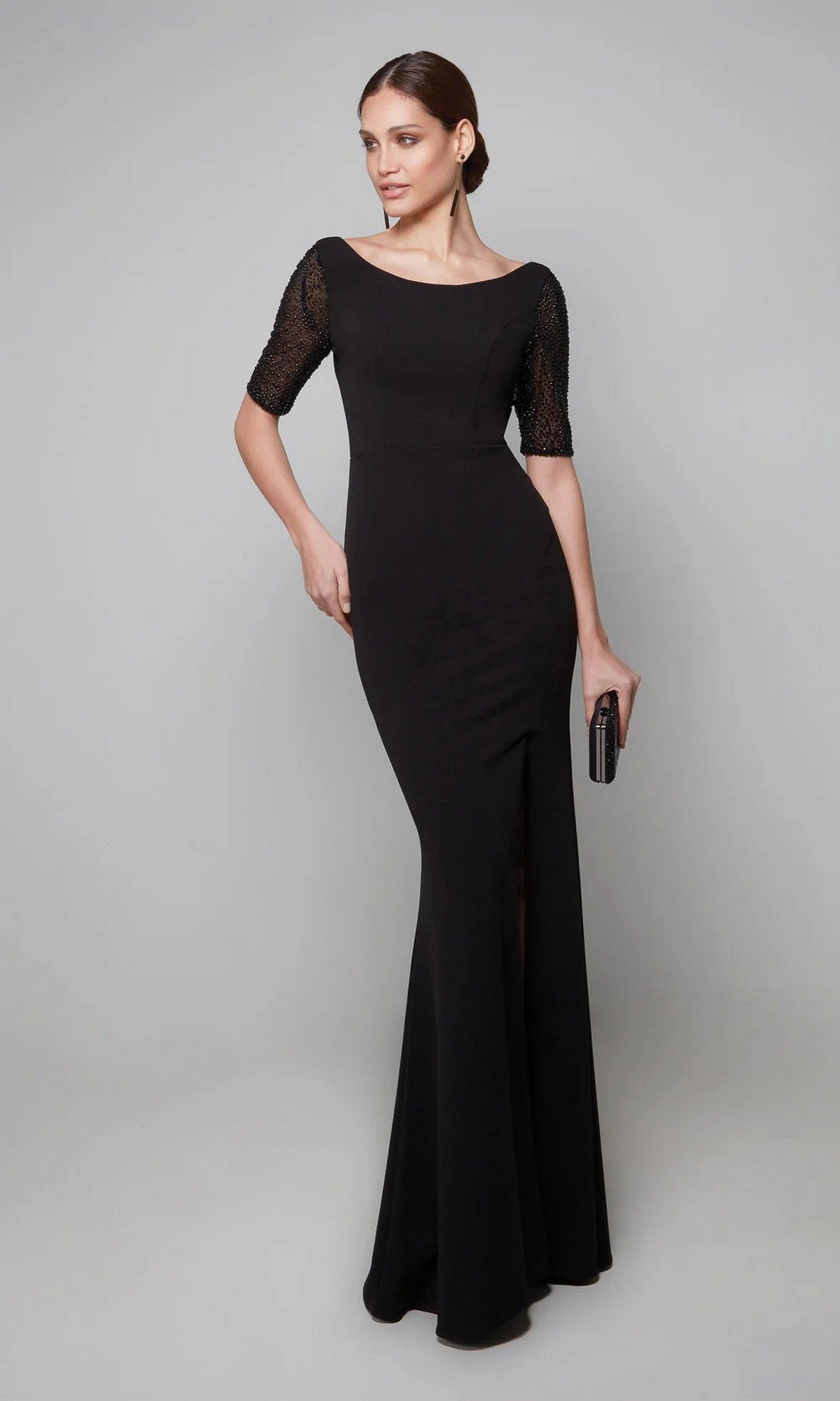 A woman in the Alyce 27628 long formal black dress with lace half-sleeves and a thigh-high slit holds a black clutch, standing against a plain light gray background.