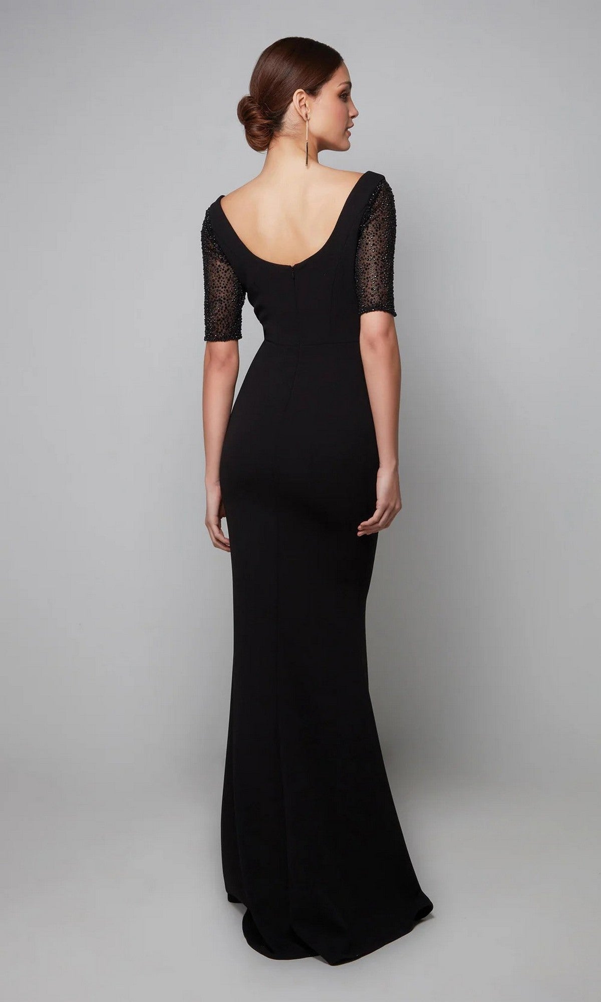 A woman faces away from the camera wearing the Alyce 27628 long formal dress—black jersey with short lace sleeves, a deep scoop back, and a thigh-high slit—set against a plain gray background.