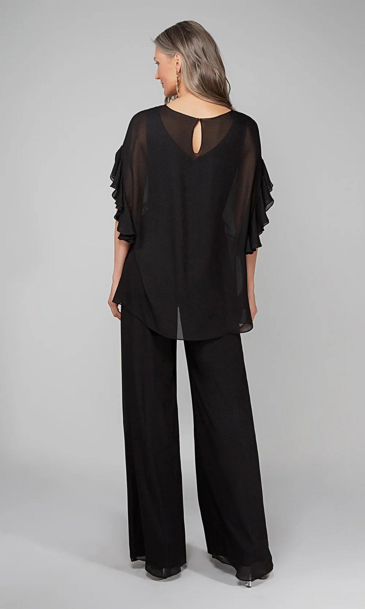 A woman faces away in the Long Formal Jumpsuit: Alyce 27633, featuring sheer ruffled sleeves and black wide-leg pants, set against a plain light gray background.