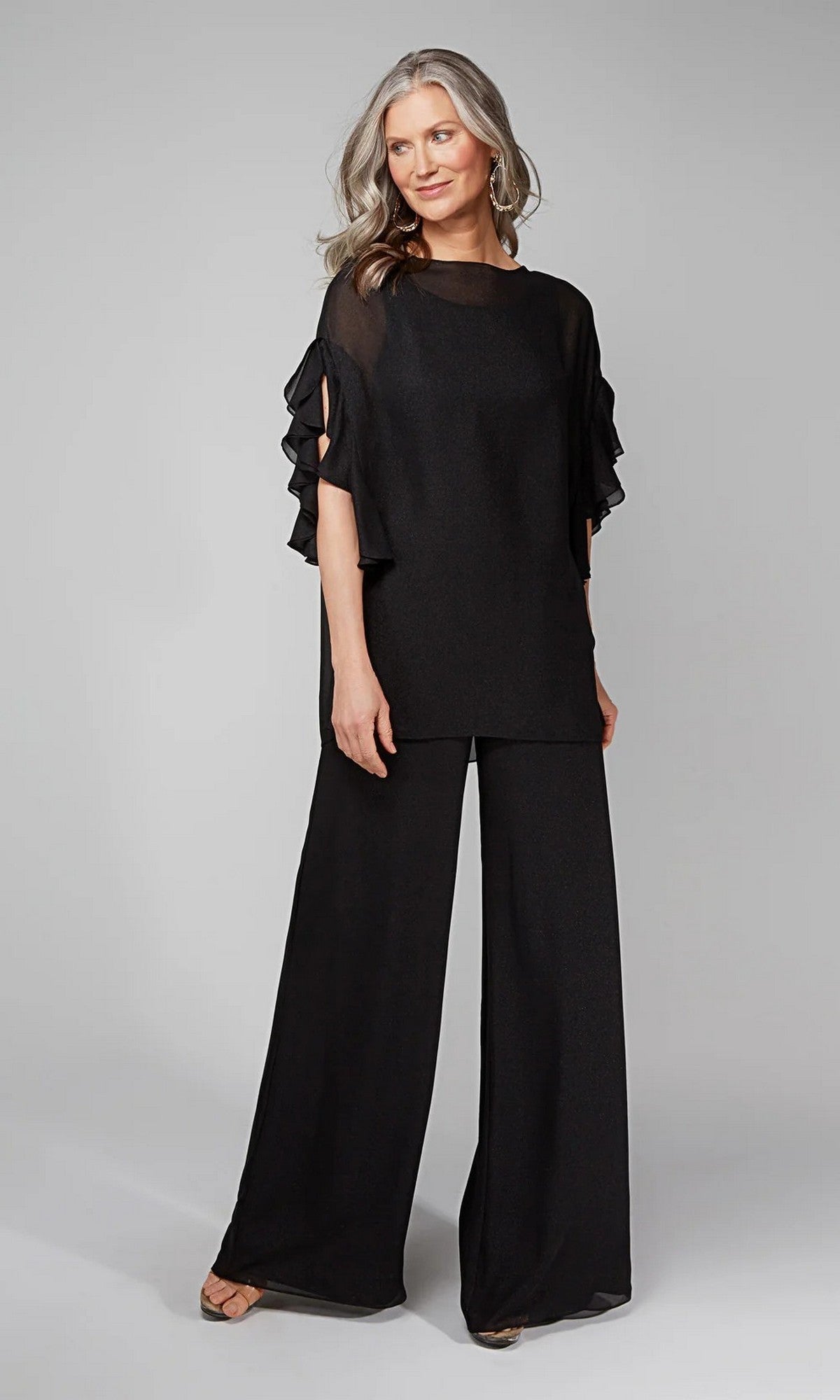 A woman with long, wavy gray hair wears the Alyce 27633 long formal jumpsuit, featuring sheer ruffled sleeves and wide-leg pants, standing against a plain gray background.