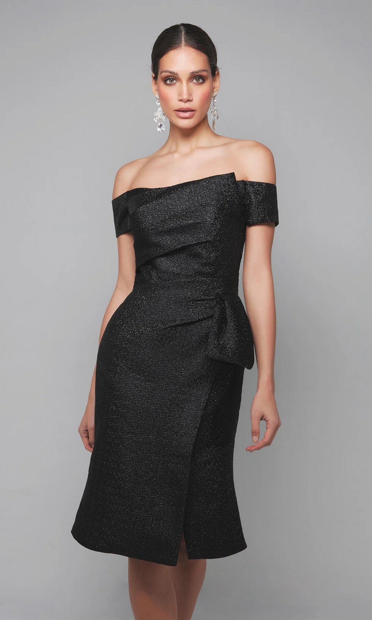 A woman models the Alyce 27638 knee-length semi-formal dress in black, featuring an off-the-shoulder neckline and side bow detail, posed against a plain gray background.