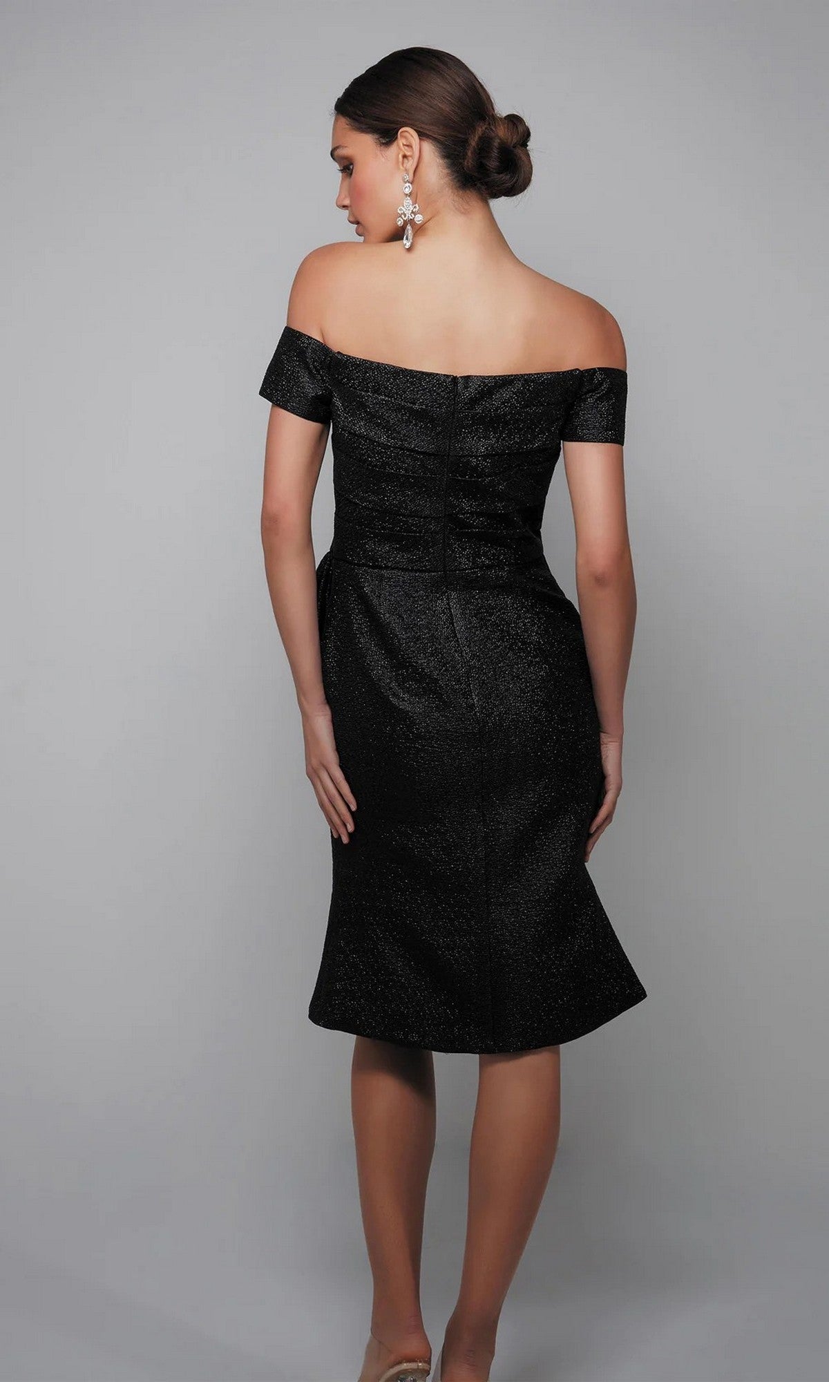 A woman poses with her back to the camera, wearing the Alyce 27638 knee-length semi-formal dress in black jacquard with an off-the-shoulder neckline, paired with dangling earrings and her hair styled in a low elegant bun.