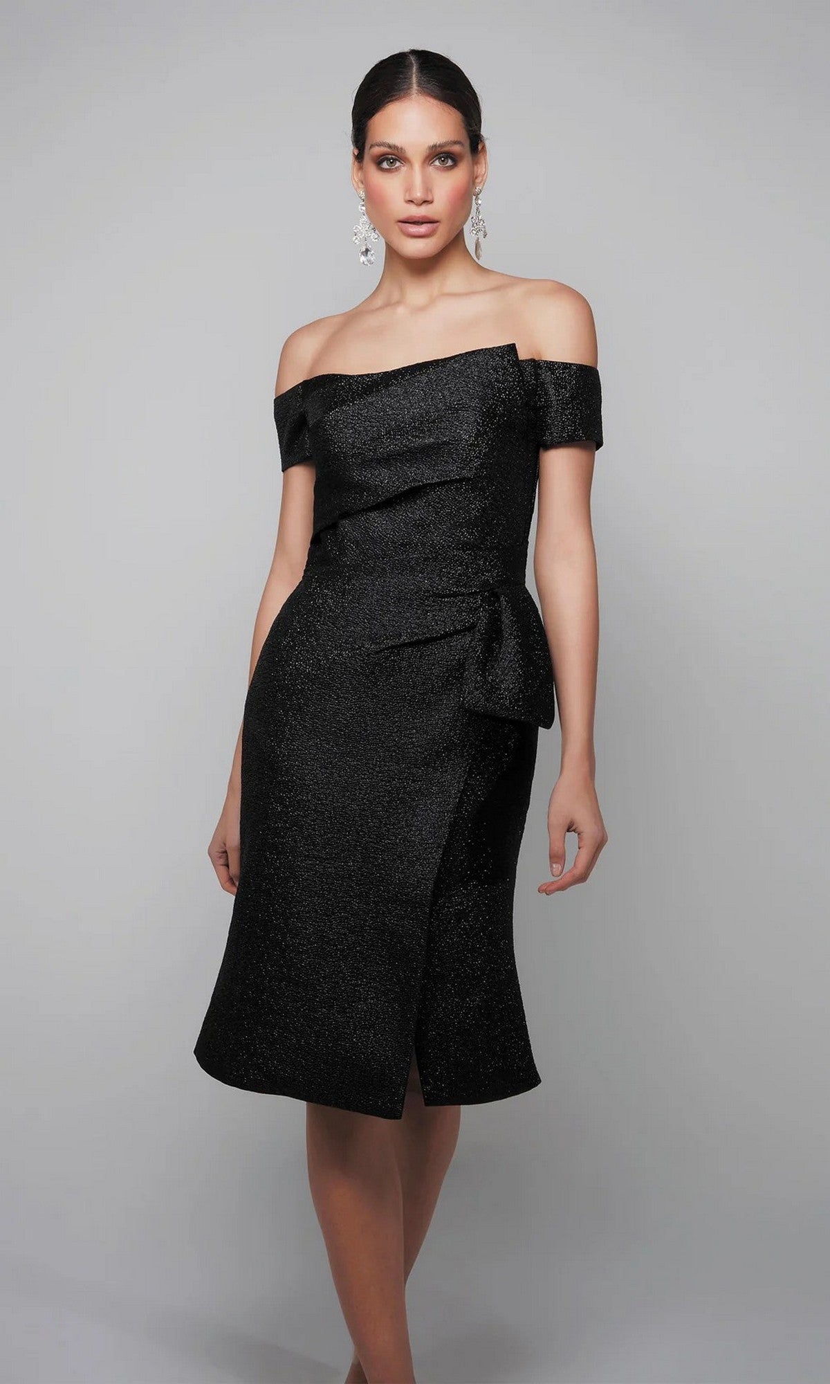 A woman poses against a plain background, wearing the Alyce 27638 knee-length semi-formal dress in black jacquard with an off-the-shoulder neckline, side bow detail, and statement earrings.