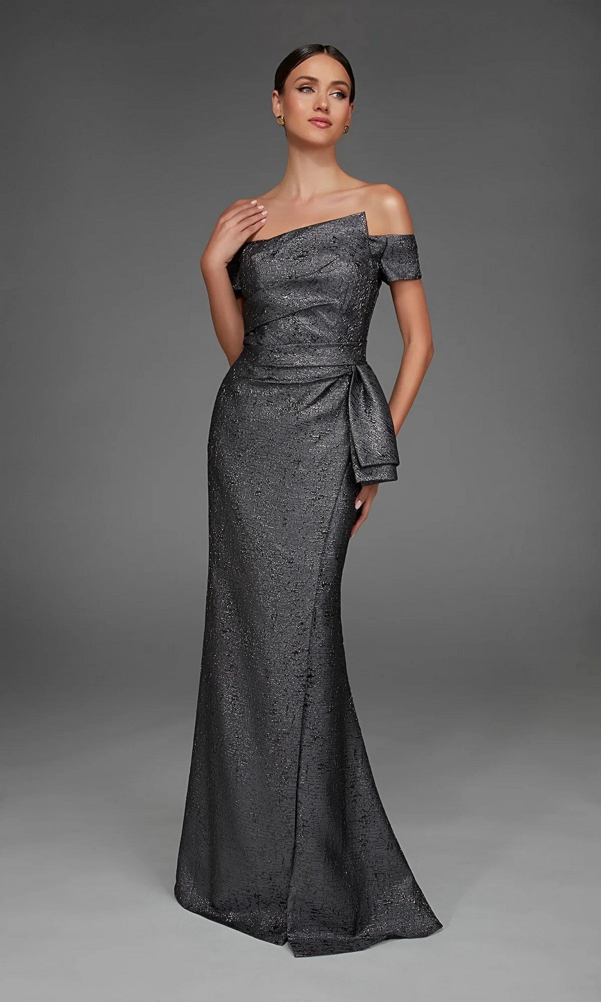 A woman in the Long Formal Dress: Alyce 27639, an off-the-shoulder, floor-length dark metallic jacquard evening gown with a side bow detail, stands against a plain gray background.