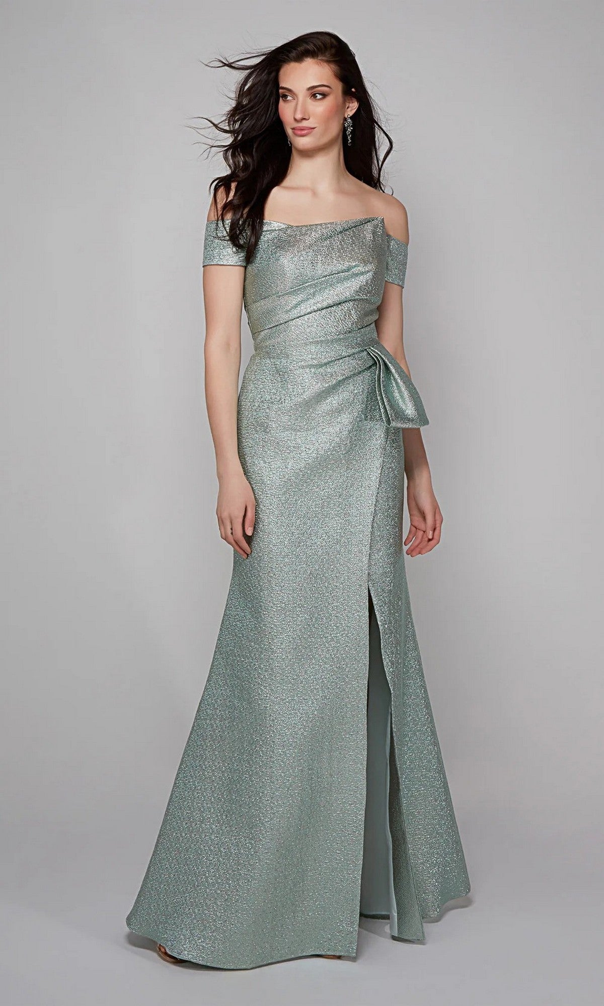 A woman wears the Alyce 27639 Long Formal Dress in Dripping Diamonds (Silver), featuring an off-the-shoulder design, fitted bodice, side bow detail, and floor-length skirt against a plain background.