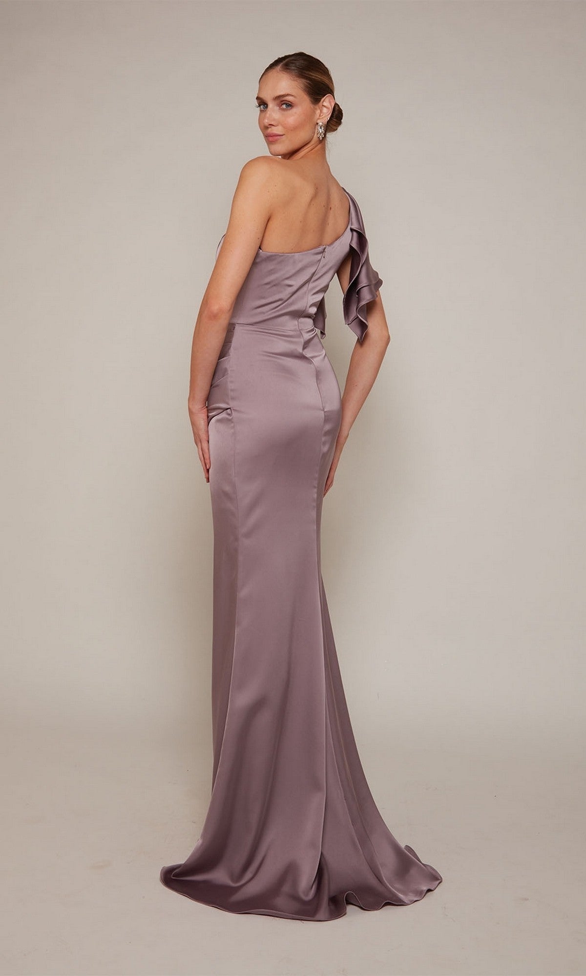 Woman wearing the Alyce 27656 long formal dress—one-shoulder satin style with shoulder ruffle and sheath faux wrap skirt—stands against a plain light background, facing away and looking over her shoulder.