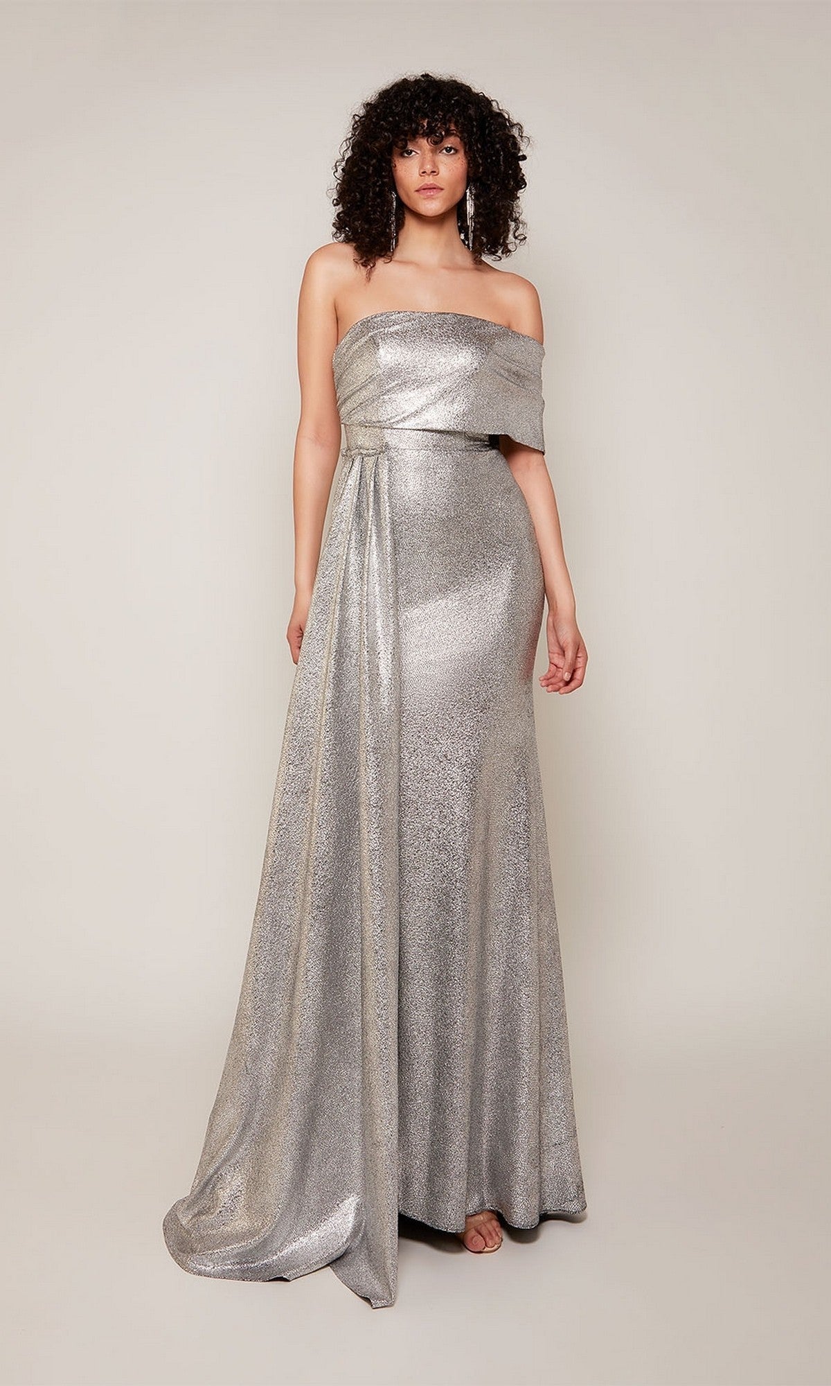 A person stands against a plain light background wearing the Alyce 27663 long formal dress, featuring a silver metallic jacquard fabric, off-the-shoulder neckline, draped detail, and a thigh-high side slit.