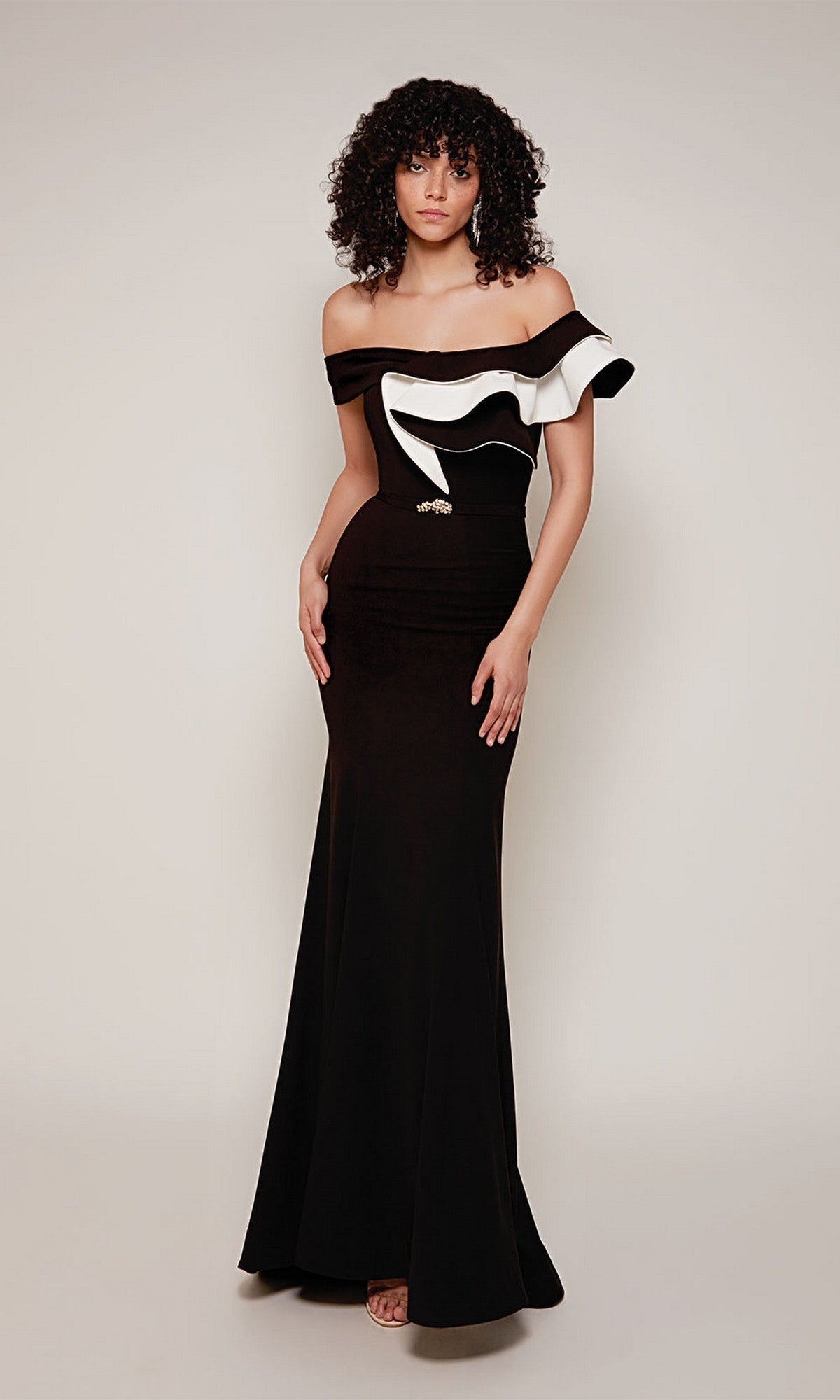 A woman wears the Alyce 27672 long formal dress in black/ivory, featuring a floor-length, off-the-shoulder sheath silhouette with two-tone ruffle detail, standing against a plain light background.