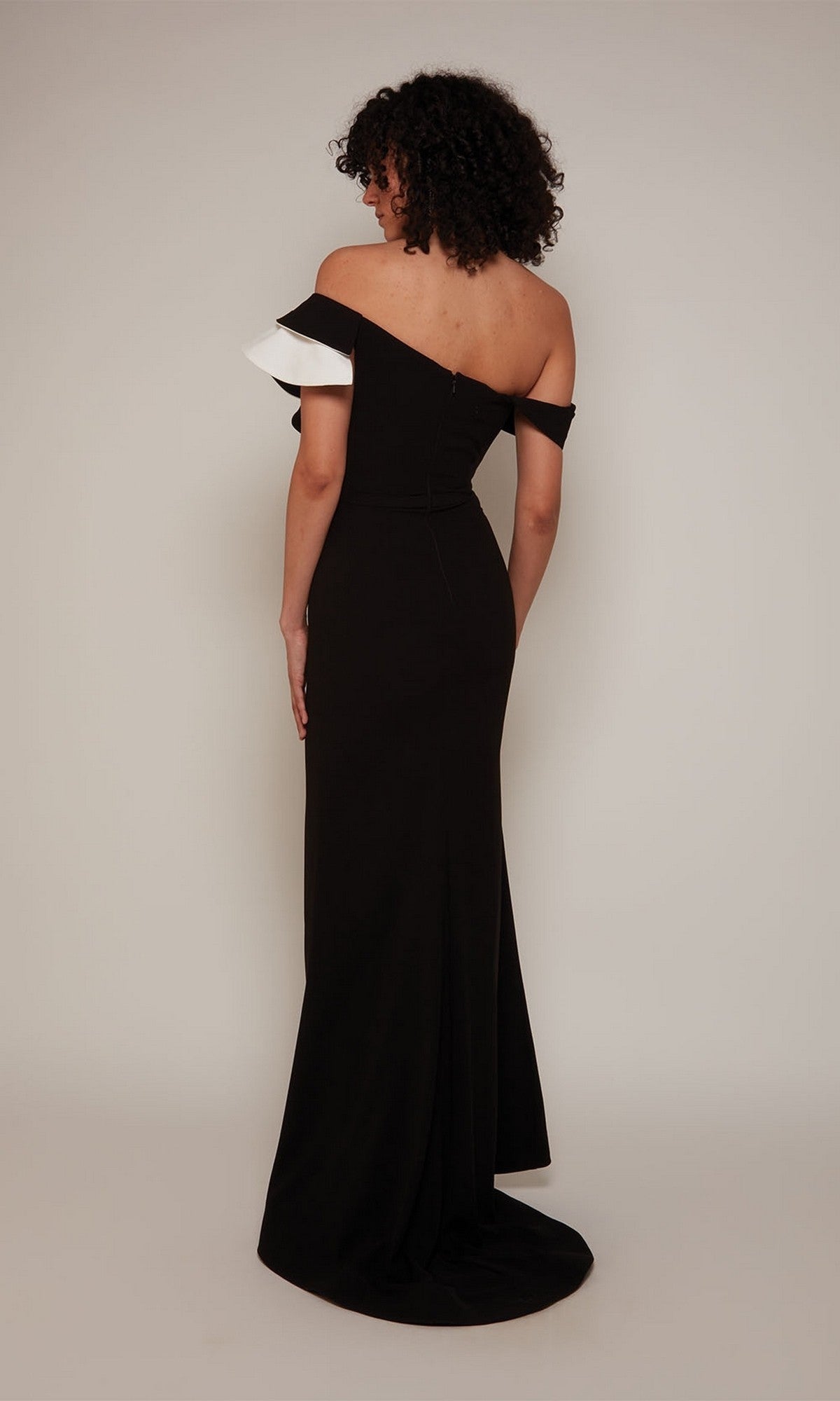 A person, facing away, wears the Alyce 27672 long formal dress—a fitted black sheath evening gown with off-the-shoulder styling and white detailing on one sleeve—posed against a plain light background.