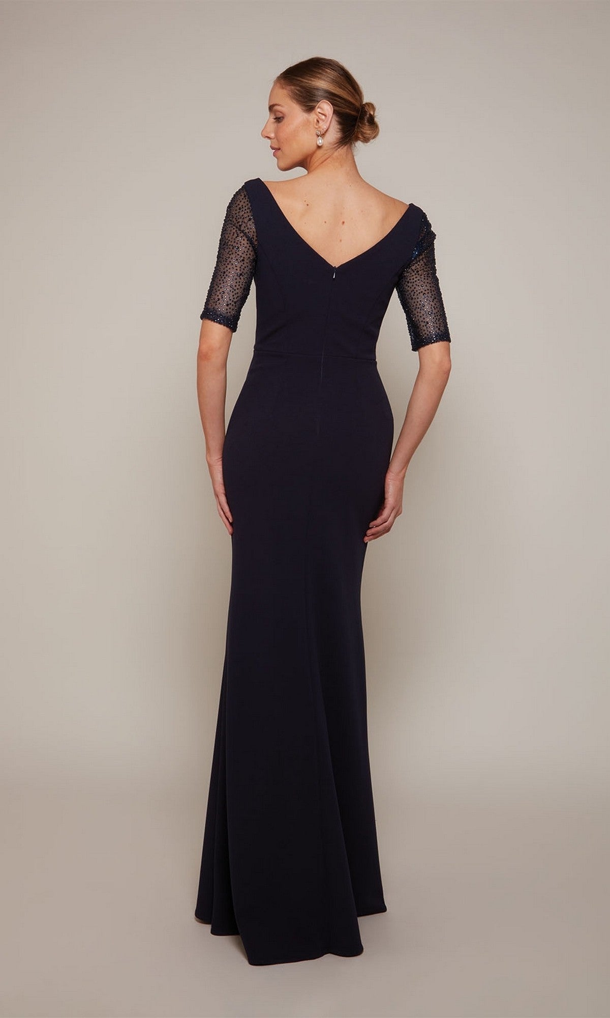 A woman stands with her back to the camera in the Alyce 27677, a floor-length dark formal gown with sheer short sleeves and deep V-back—ideal for black-tie events or as a mother-of-the-bride dress.