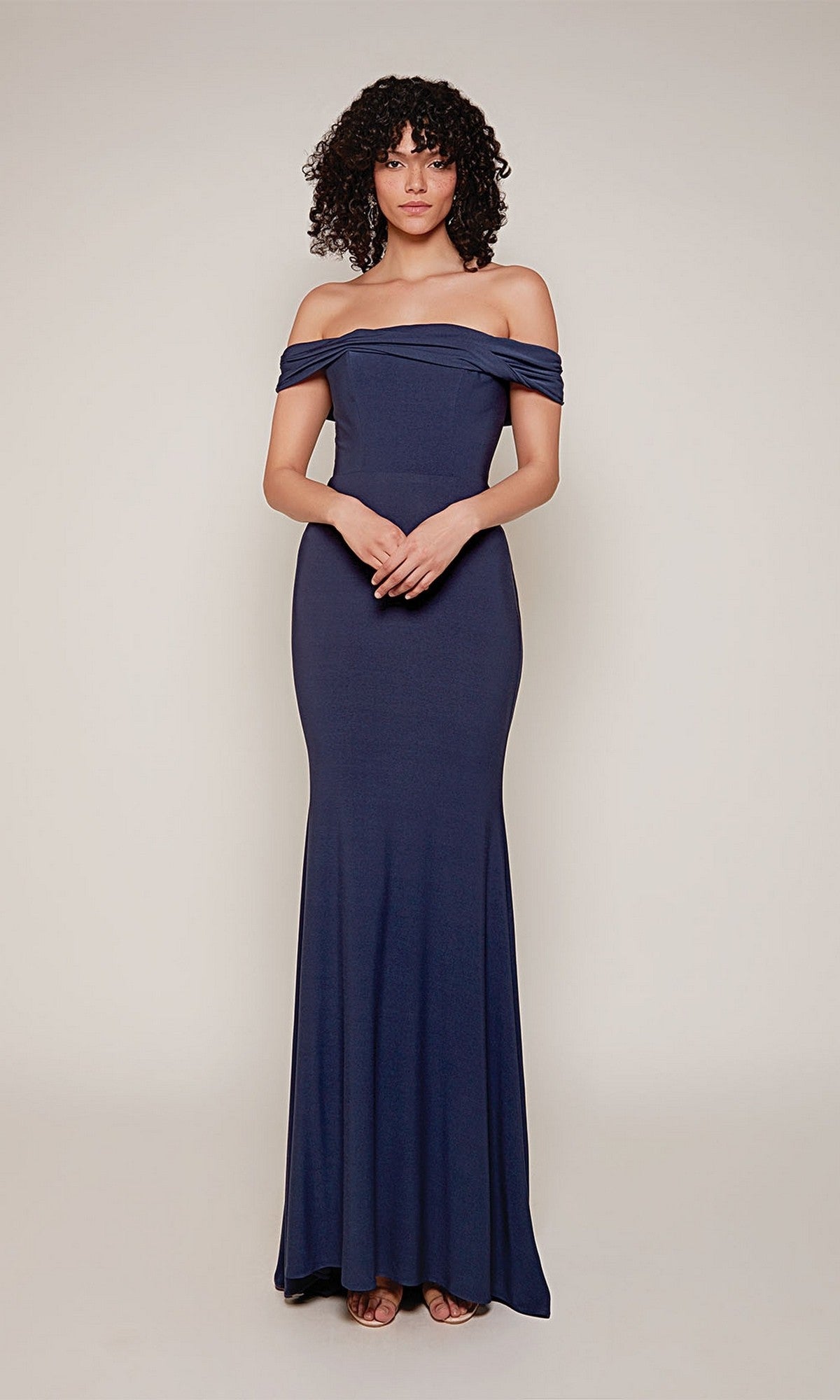 A person stands against a plain background, wearing the Alyce 27680 long formal dress in navy—an off-the-shoulder, slim-fit, floor-length gown—with hands clasped in front.