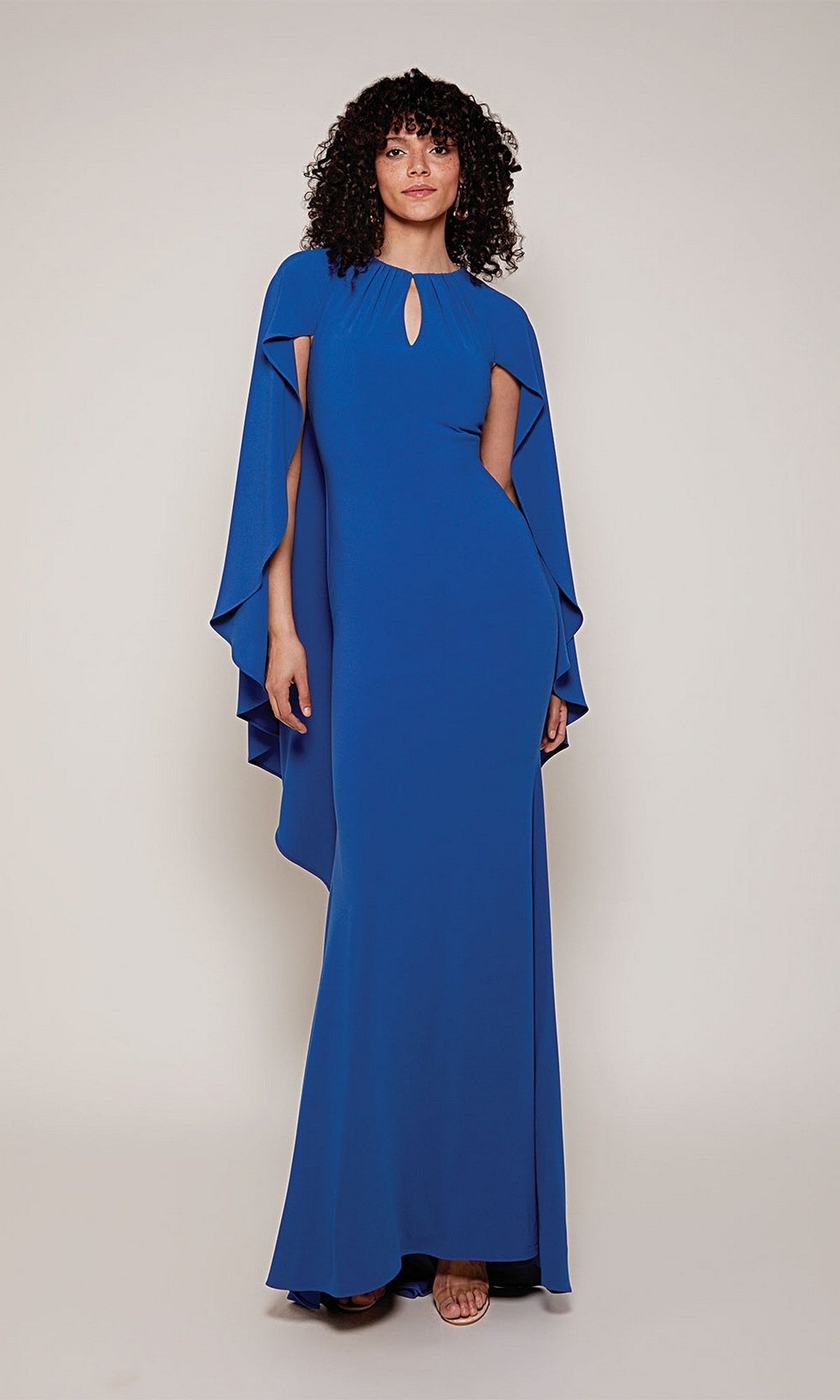 A woman models the Alyce 27683 long formal dress in royal, featuring a floor-length column silhouette, keyhole neckline, and draped high-low cape sleeves, set against a plain light background.