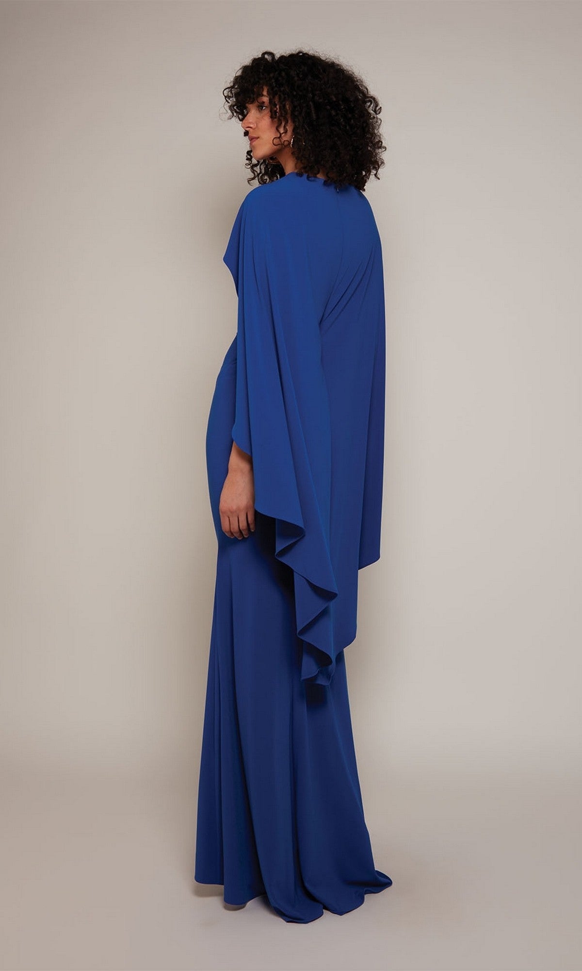 A person with curly hair stands sideways, wearing the Alyce 27683 long formal column dress featuring flowing high-low cape sleeves, set against a plain light background.