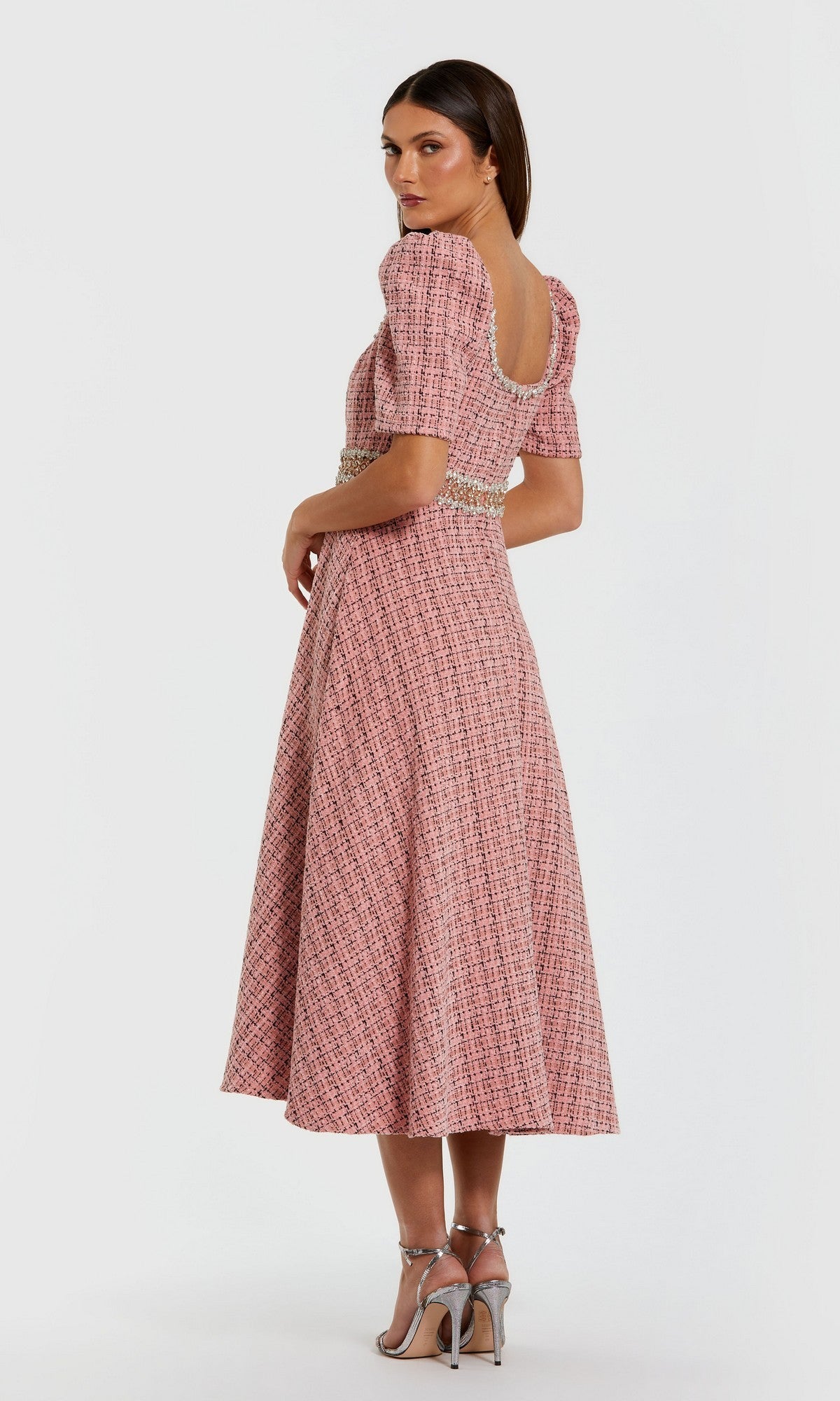 A woman in profile wears the Mac Duggal 27687 Midi Wedding Guest Dress—pink and white with puff sleeves, open back, rhinestone-beaded belt—paired with silver high heels, set against a plain background.