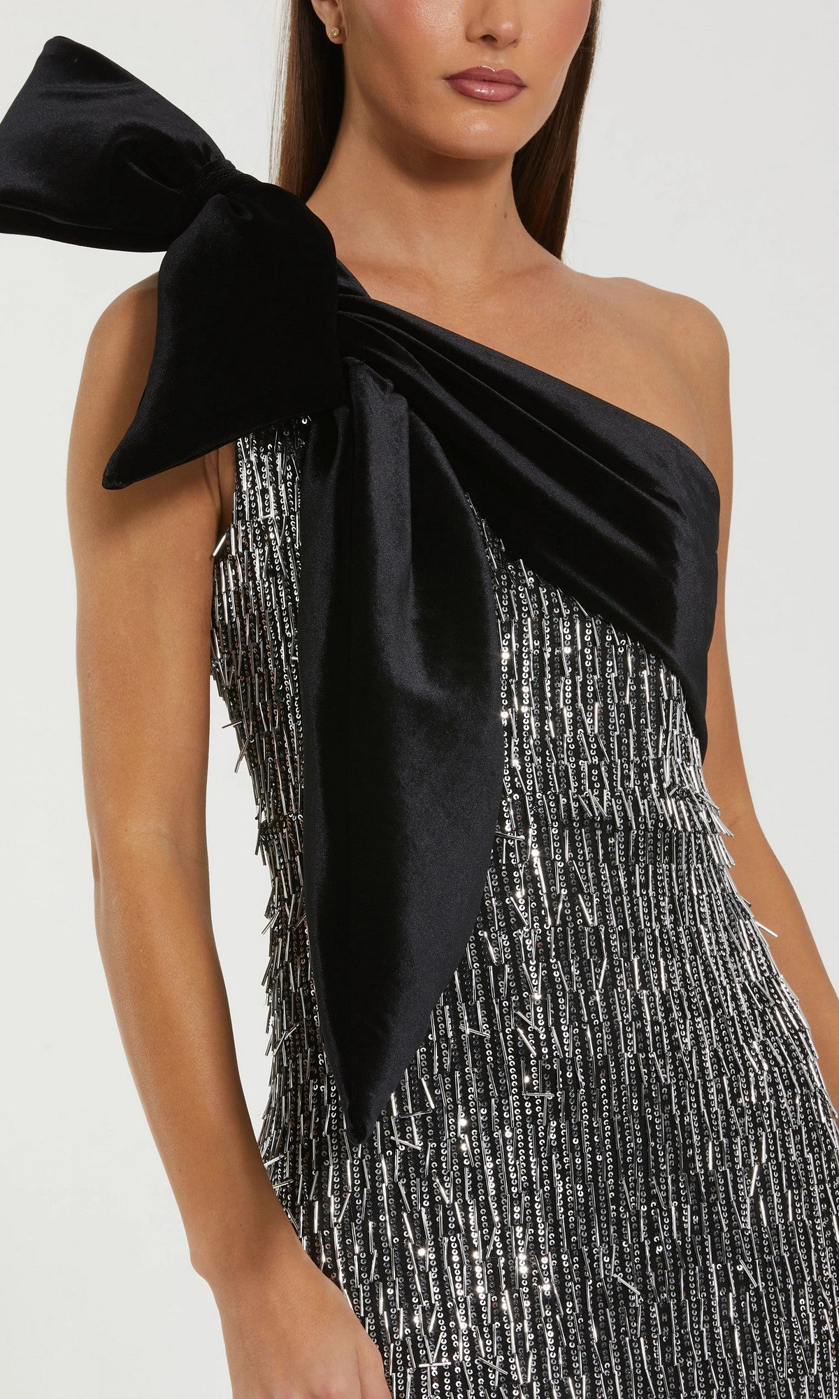 A woman wears the Mac Duggal 27690 long formal dress, a one-shoulder sequin gown with a large black velvet bow and textured silver and black fabric.