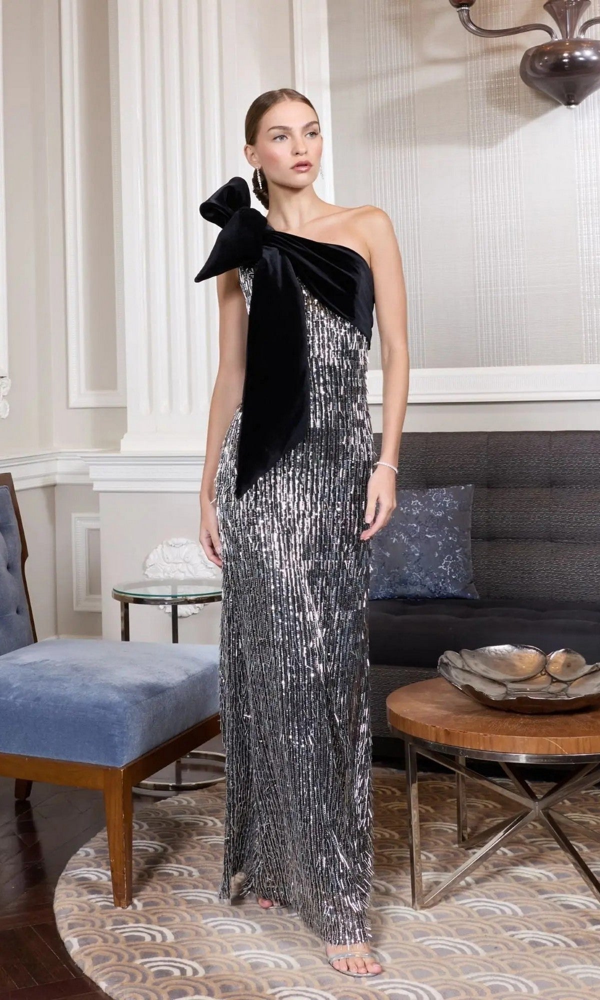 A woman stands in a stylish living room wearing the Mac Duggal 27690 long formal dress in black silver, featuring a one-shoulder design with sequins and a large bow.