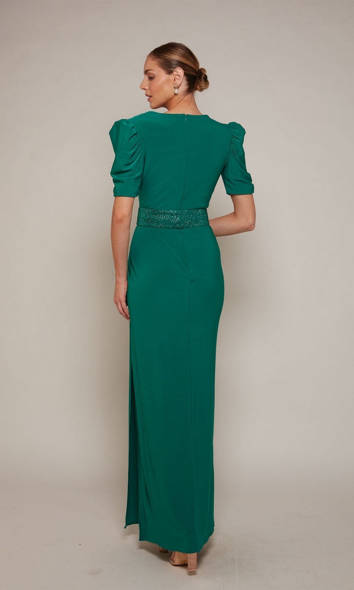 A woman with her hair in a low bun faces away, wearing the Alyce 27690 emerald long formal dress featuring a v-neckline, short puffed sleeves, and a beaded waist belt.